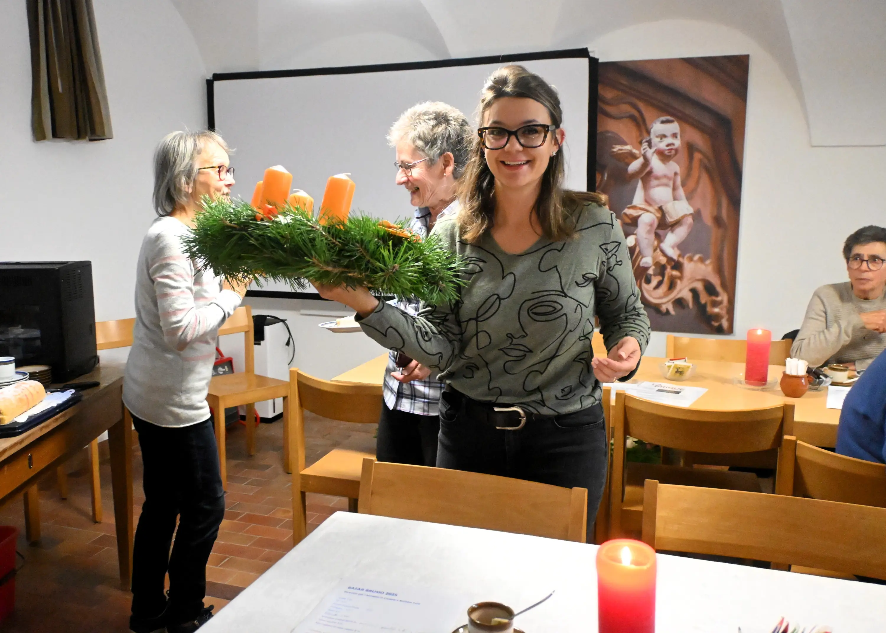 Una giovane donna sorride mentre tiene in mano un centrotavola di Natale con candele arancioni. Sullo sfondo, altre persone chiacchierano in un ambiente accogliente.