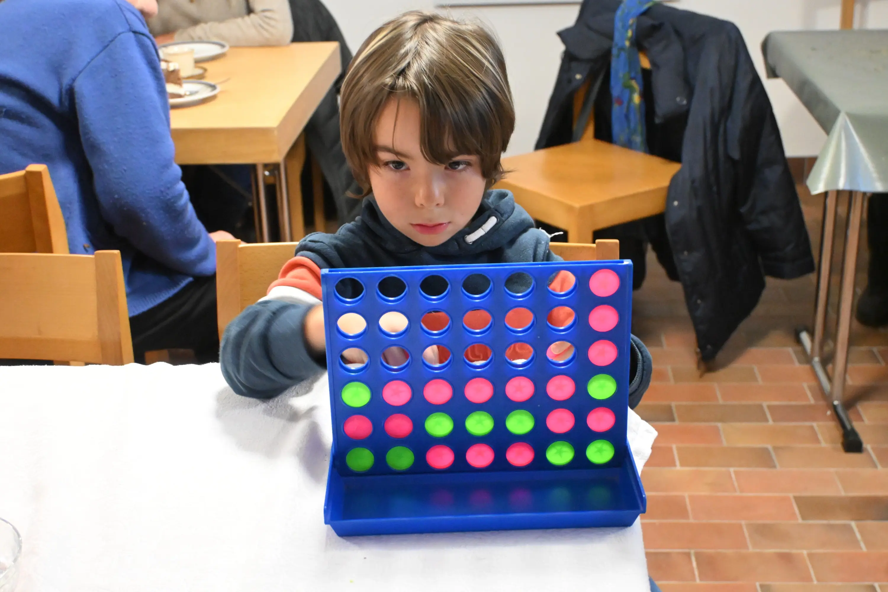 Un ragazzo concentra mentre gioca a Forza Quattro, posizionando un token in una griglia blu con fori rossi e verdi. Sullo sfondo, si vedono altre persone sedute a tavoli di legno.