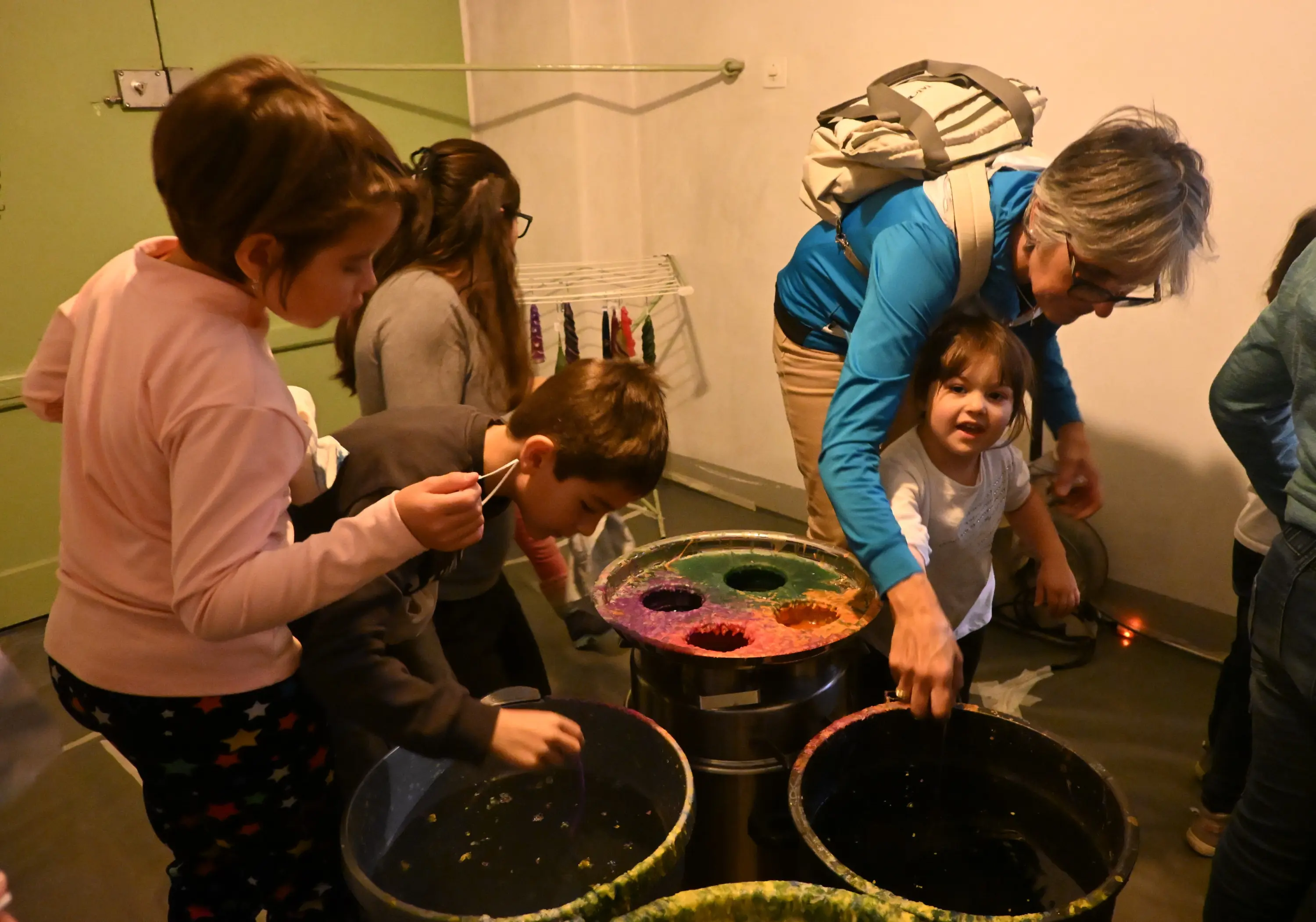 Un gruppo di bambini e un'adulta si trovano in una stanza, mentre eseguono attività di pittura. Alcuni utilizzano dei secchi con colori diversi, mentre uno dei bambini guarda sorridendo verso l'obiettivo.