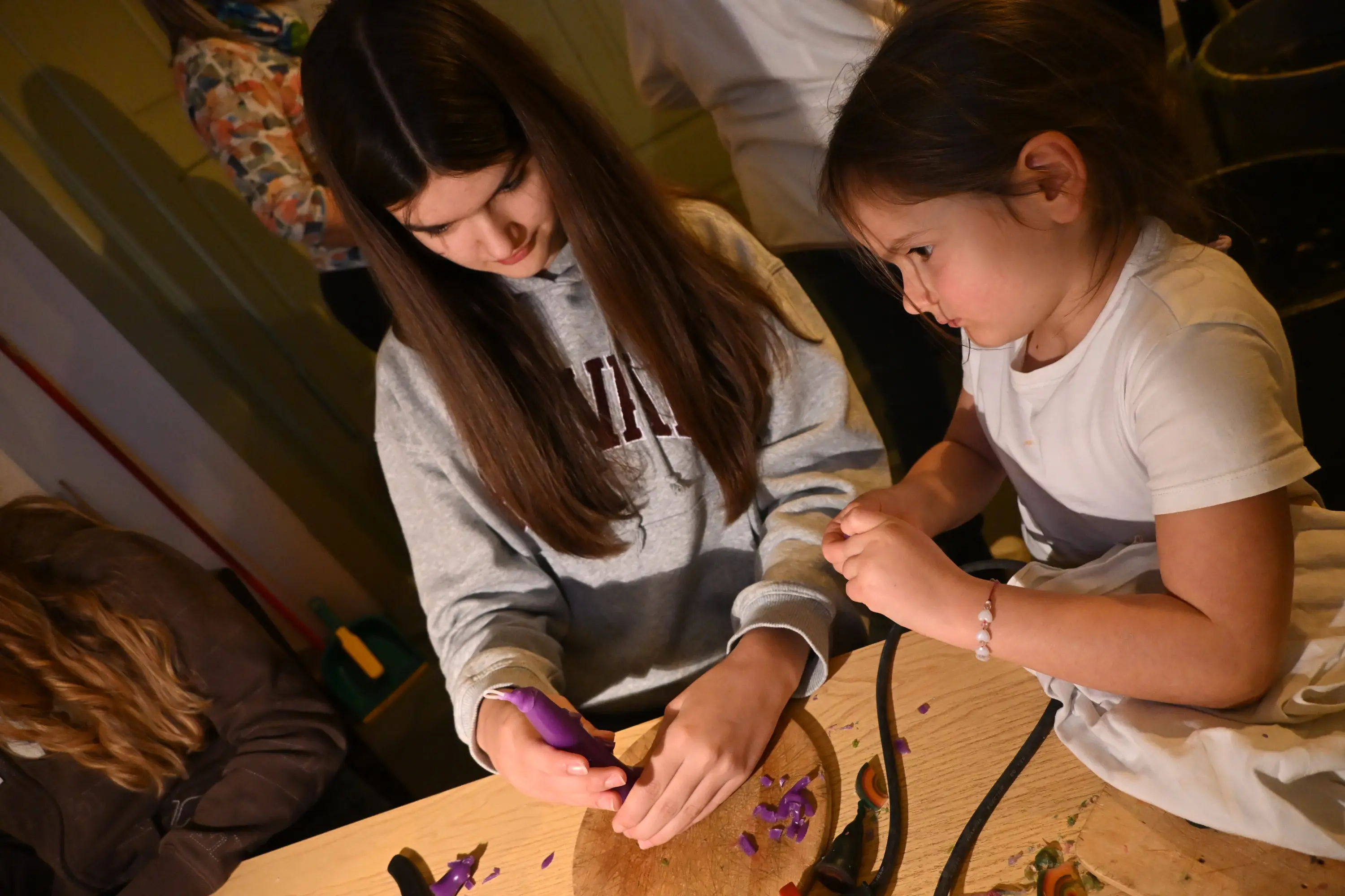 Due ragazze stanno creando oggetti artigianali su un tavolo. L'adolescente, con i capelli lunghi e castani, usa un attrezzo per modellare un materiale, mentre la più piccola osserva attentamente e tiene in mano dei pezzi colorati.