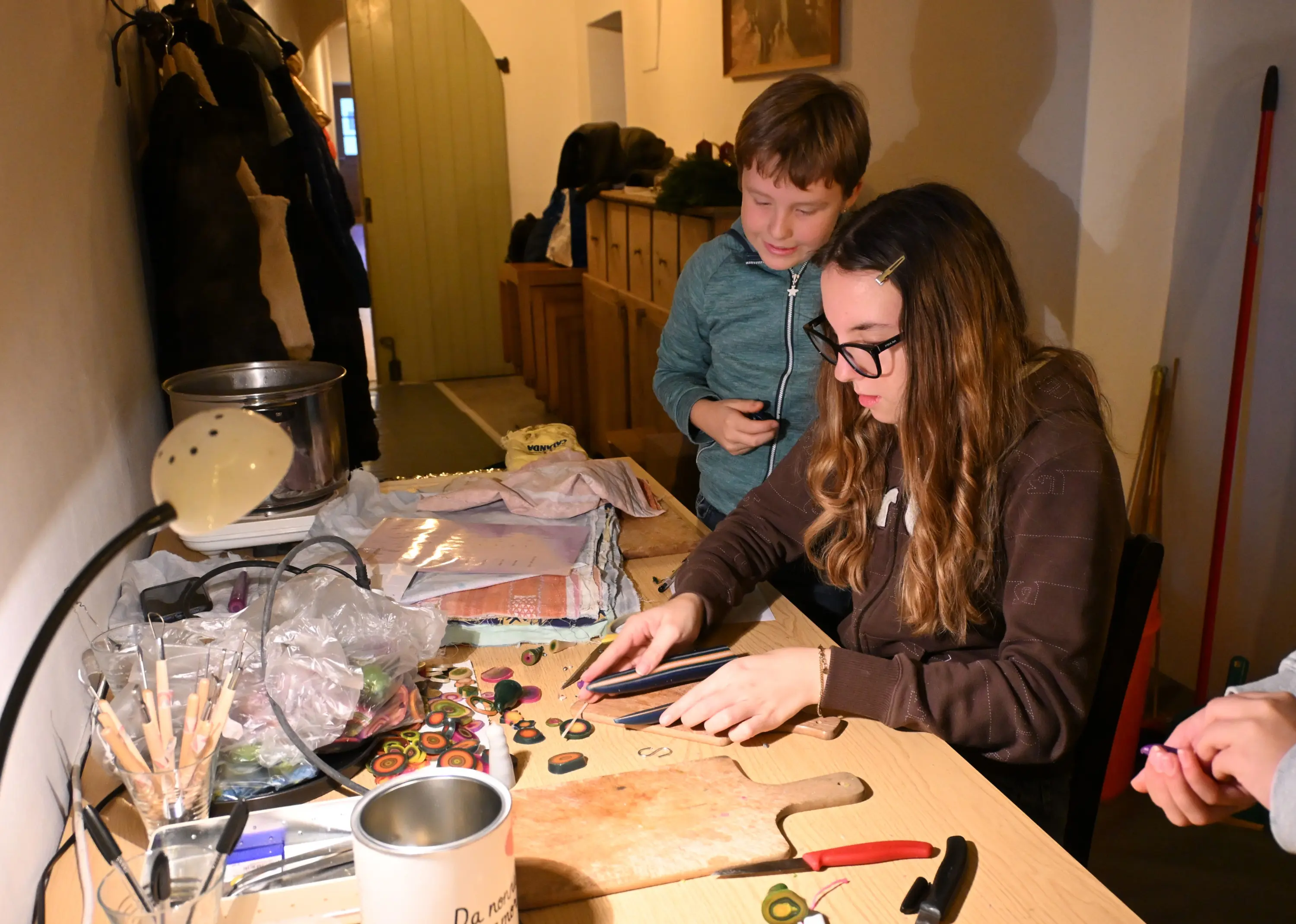 Due ragazzi lavorano a un tavolo, creando oggetti artigianali. Una ragazza sta tagliando materiali con attenzione, mentre un ragazzo osserva con interesse. Sul tavolo ci sono vari materiali e attrezzi per il fai-da-te.