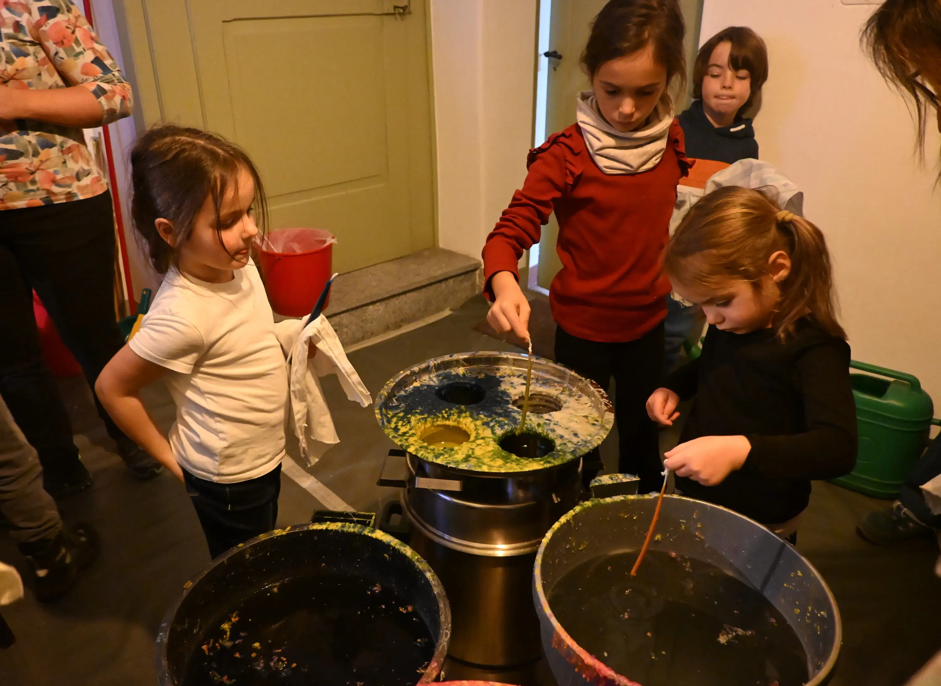 Due bambine stanno mescolando colori in grandi contenitori. Una delle ragazze usa una bacchetta mentre l'altra osserva attentamente. Sullo sfondo, un bambino e un adulto guardano.