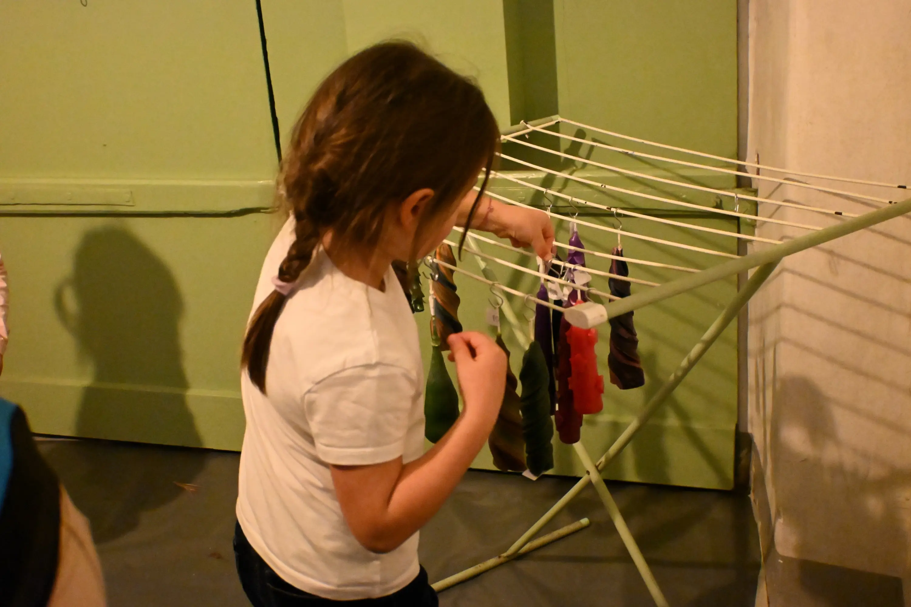Una bambina con trecce osserva e tocca delle candele colorate appese a uno stendibiancheria in una stanza con pareti chiare.