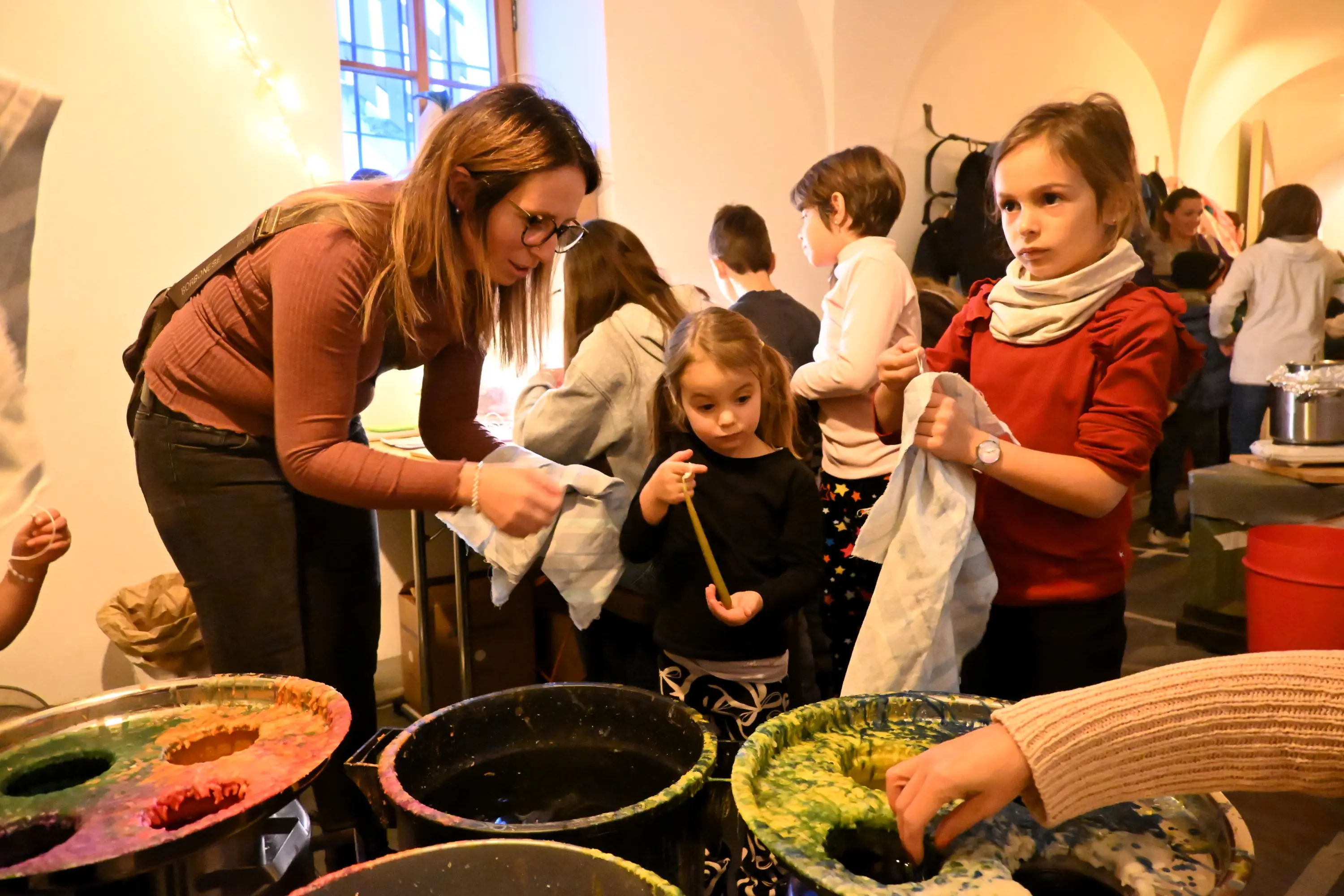 Una donna aiuta due bambine a colorare tessuti in un laboratorio creativo. Diversi vasi con colori vivaci sono visibili sul tavolo, mentre altri bambini partecipano attorno. L'atmosfera è vivace e collaborativa.