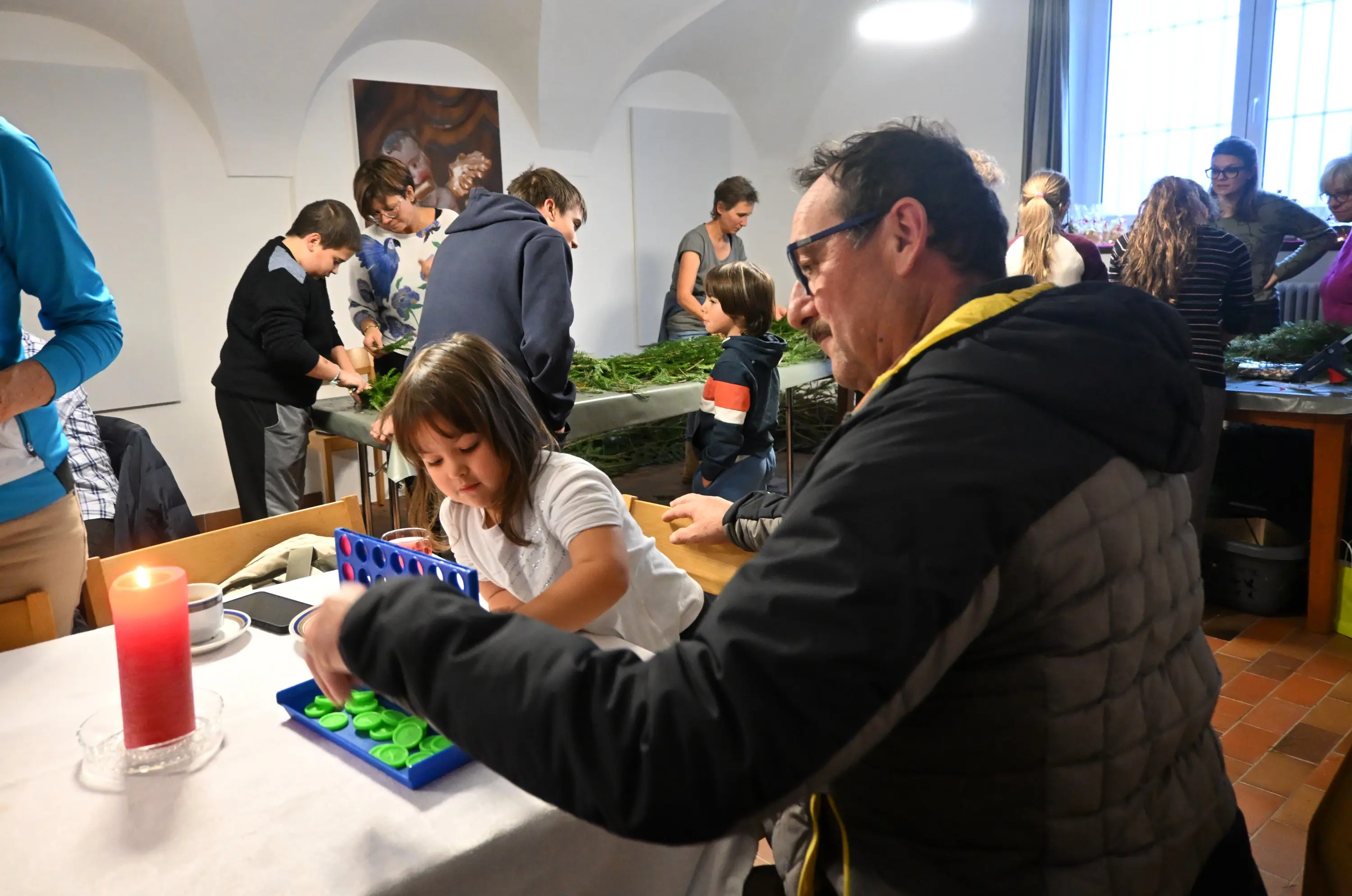 Un gruppo di persone di diverse età si riunisce in una sala, impegnate in attività manuali. Un uomo adulto gioca con una bambina, mentre altri partecipano a lavori creativi sullo sfondo. Una candela accesa è presente su un tavolo.