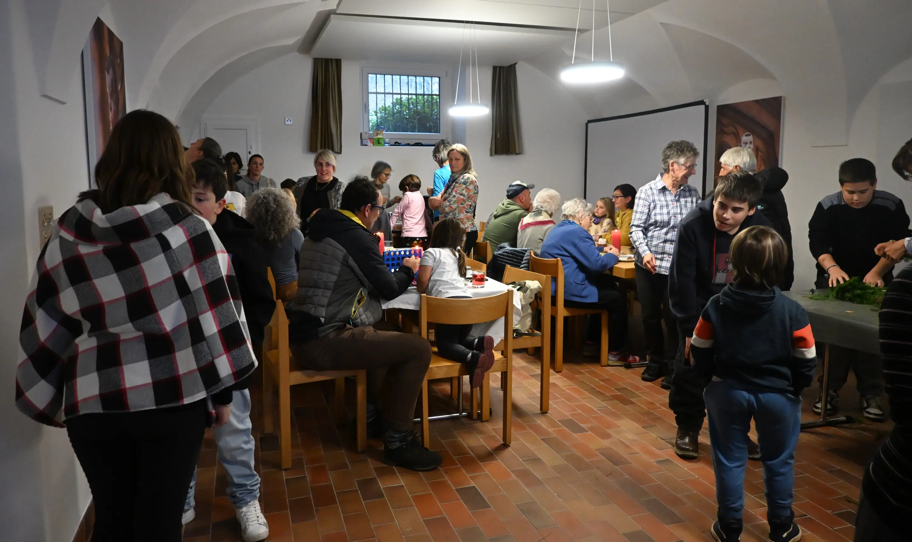 Una folla di persone riunite in una sala utilizzando tavoli e sedie. Alcuni bambini giocano, mentre gli adulti socializzano e chiacchierano. L'ambiente è luminoso e accogliente, con decorazioni sulle pareti.