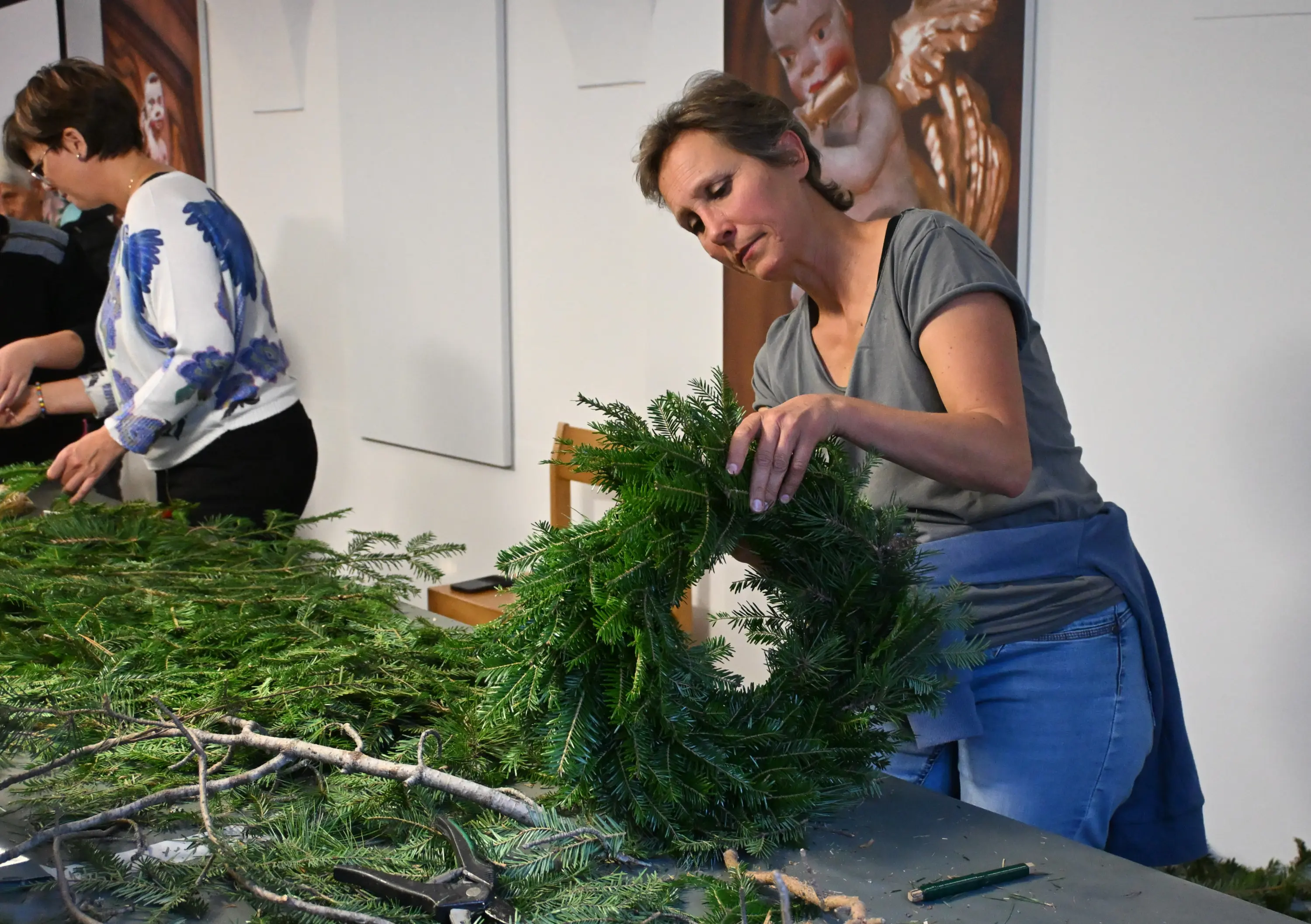 Una donna prepara una corona di rami di abete su un tavolo, con altre persone che lavorano sullo sfondo.
