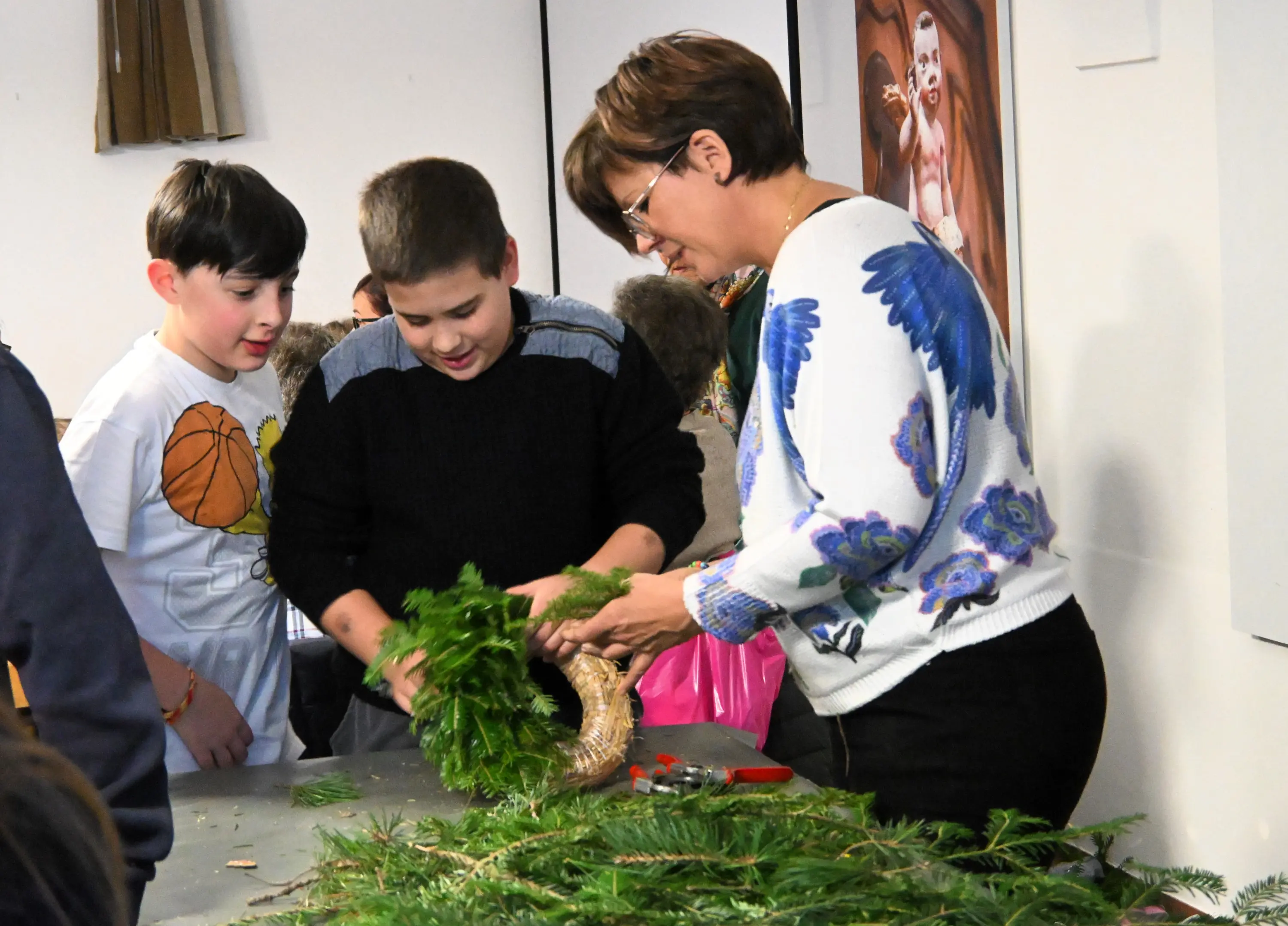 Due ragazzi osservano mentre un'insegnante crea un oggetto decorativo con rami verdi su un tavolo, circondati da materiali e attrezzi per l'artigianato.