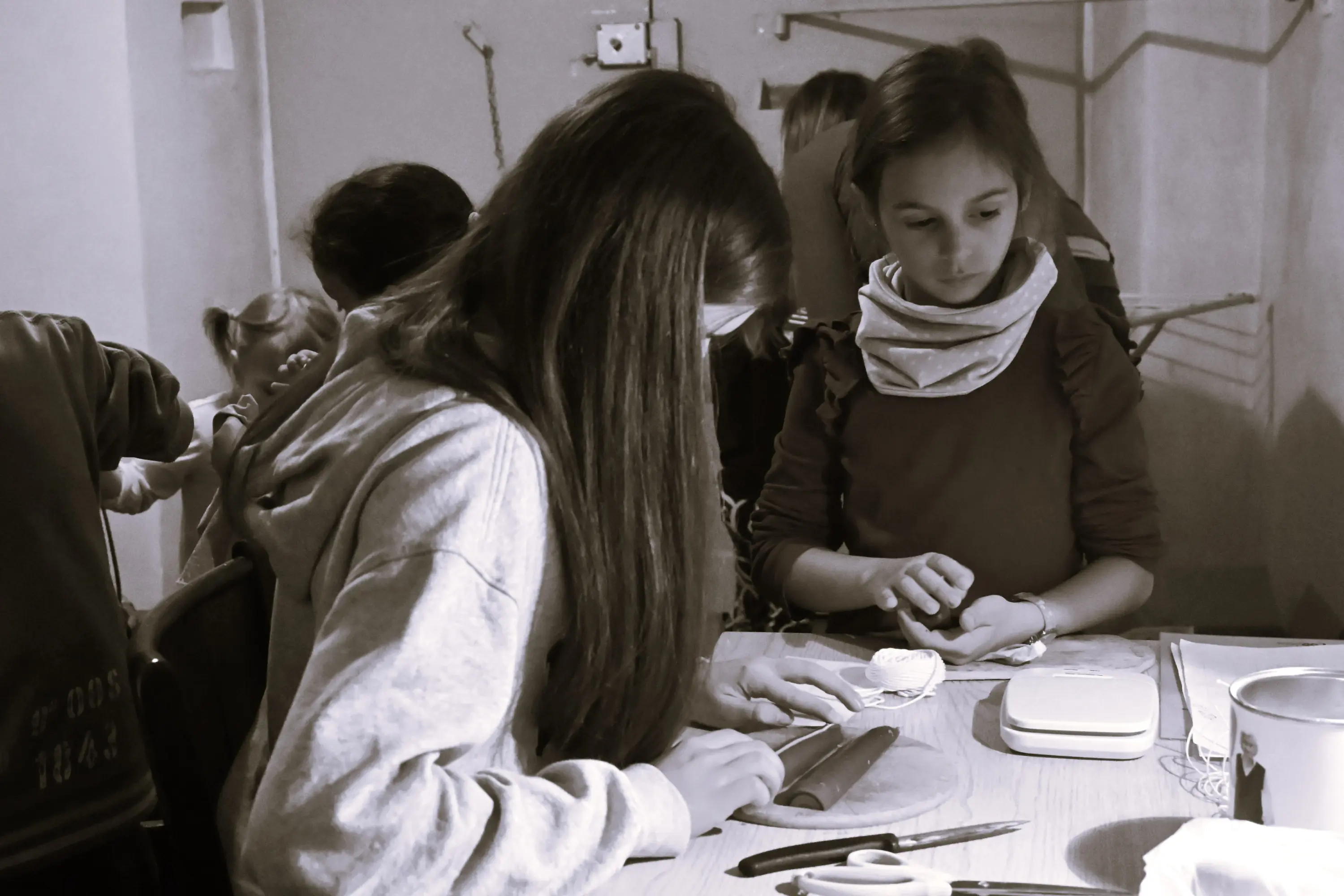 Due ragazze stanno lavorando su un progetto artigianale a un tavolo. Una ragazza ha i capelli lunghi e indossa un felpa grigia, mentre l'altra ha i capelli corti e indossa una sciarpa. Sul tavolo ci sono strumenti e materiali per