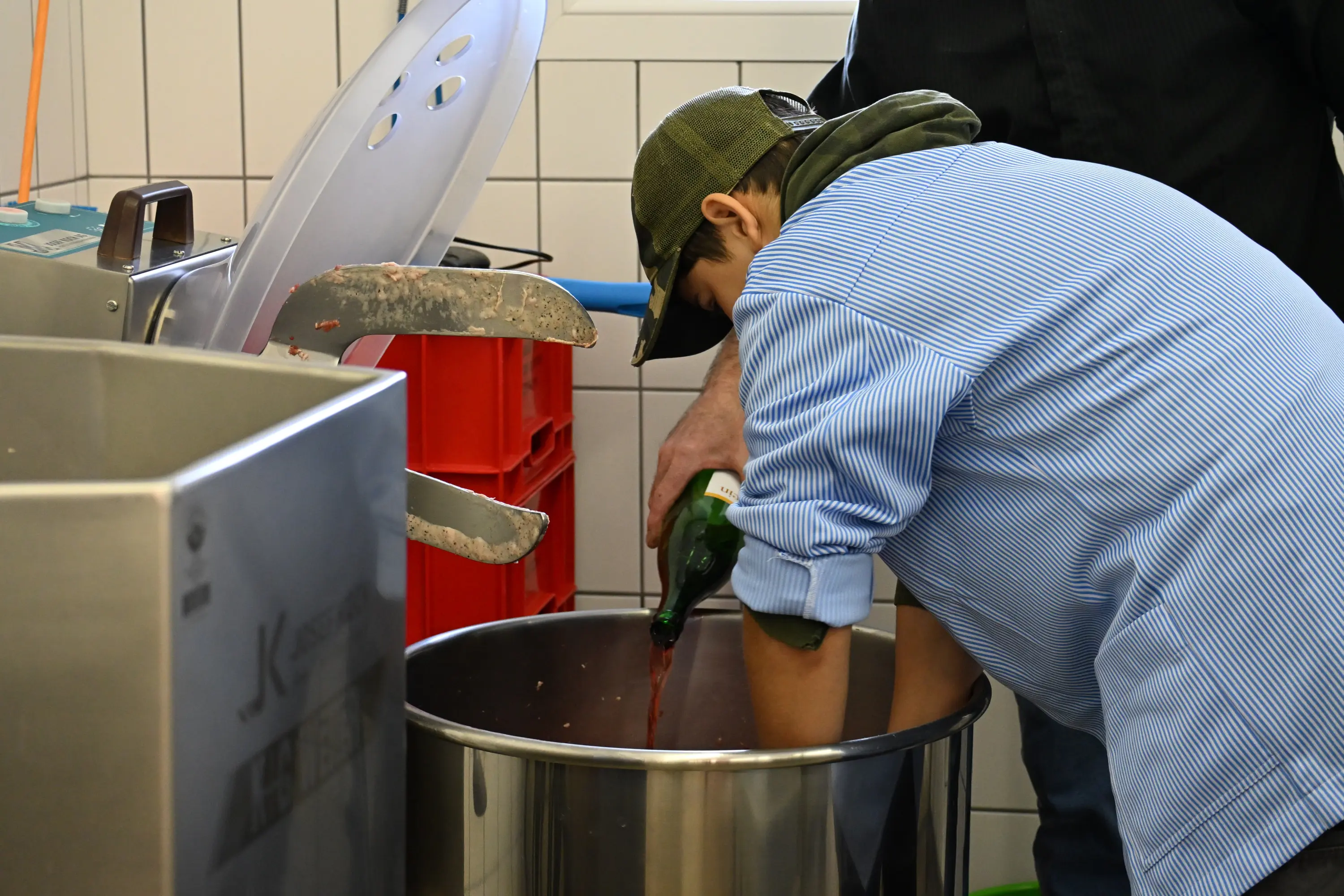 Un giovane indossa una camicia a righe e un cappellino mentre versa del vino rosso in un grande recipiente metallico. Un adulto lo assiste, con una mano che mostra un attrezzo di cucina. L'ambiente è di un laboratorio alimentare.