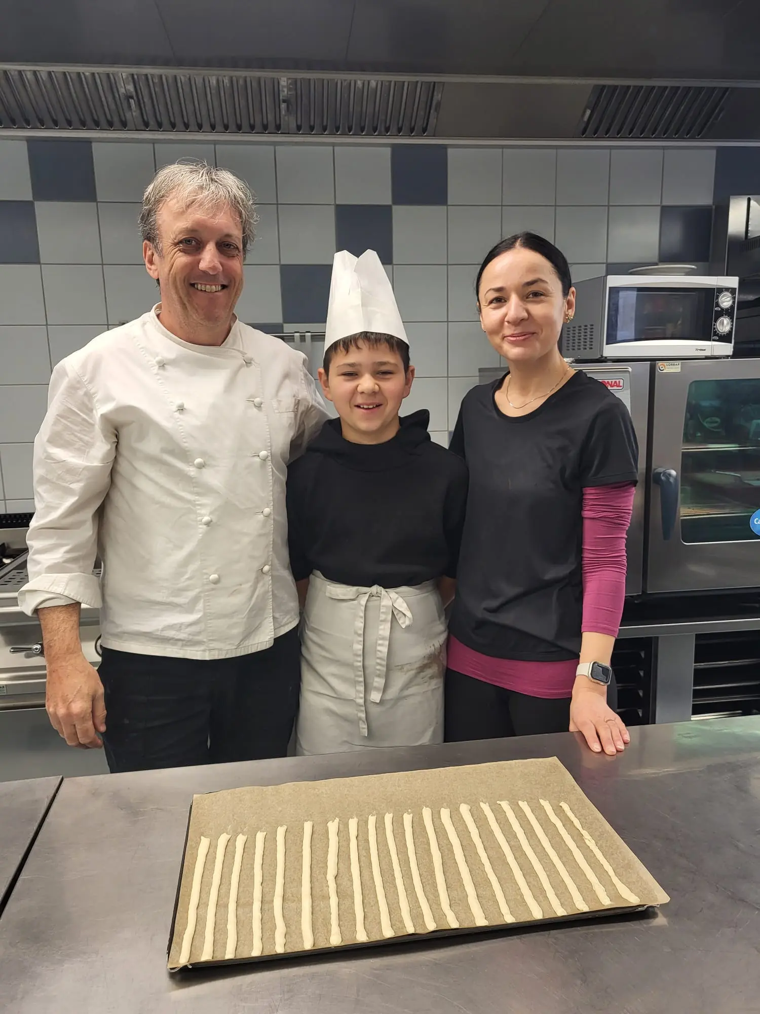 Tre persone sorridono in una cucina professionale. Un chef adulto indossa un'uniforme bianca e un cappello da cuoco, mentre un ragazzo con un cappello da cuoco e un grembiule è al centro. Una donna con una maglietta nera si