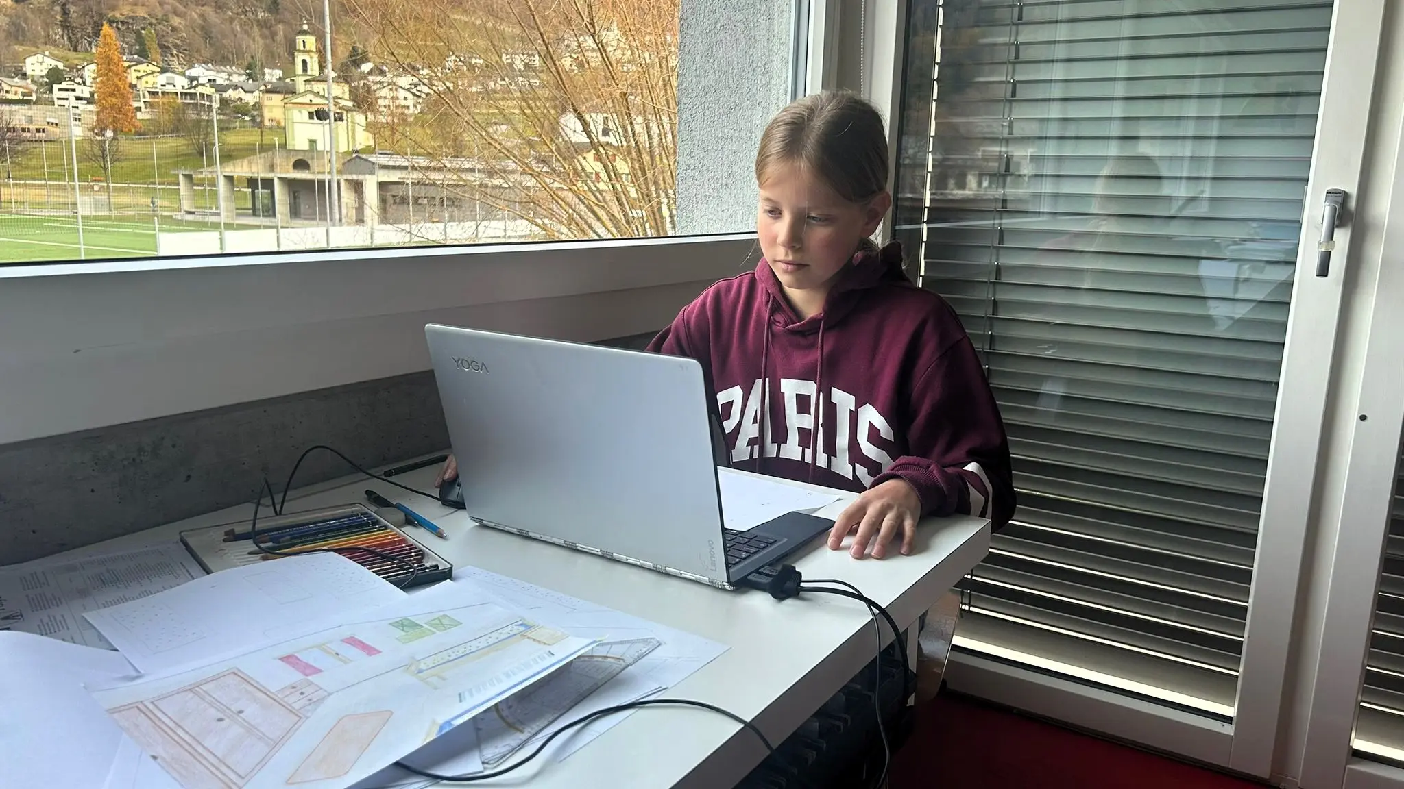 Ragazza seduta a una scrivania con un computer portatile, immersa nello studio. Circondata da fogli di carta e materiale da disegno, con una finestra che mostra un paesaggio esterno.