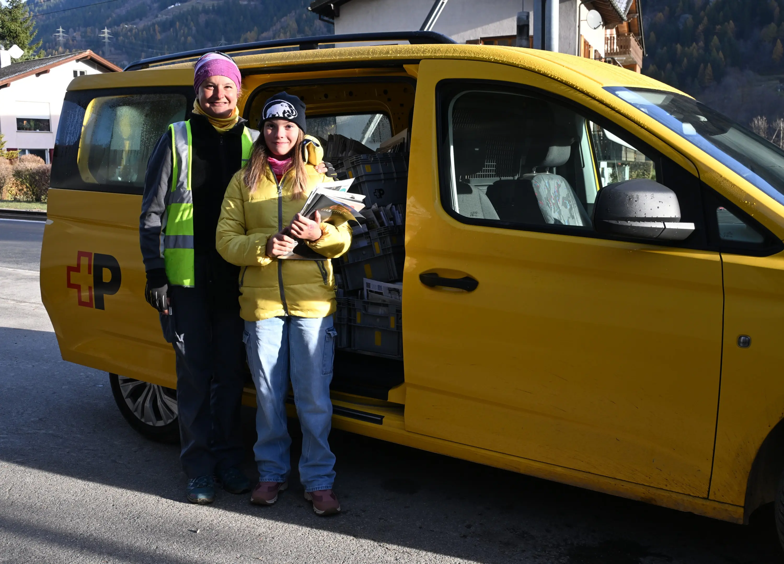 Due persone sorridenti, una con gilet riflettente, posano accanto a un furgone giallo con porte aperte, mostrando scatole di materiali all’interno. Un paesaggio montano si intravede sullo sfondo.