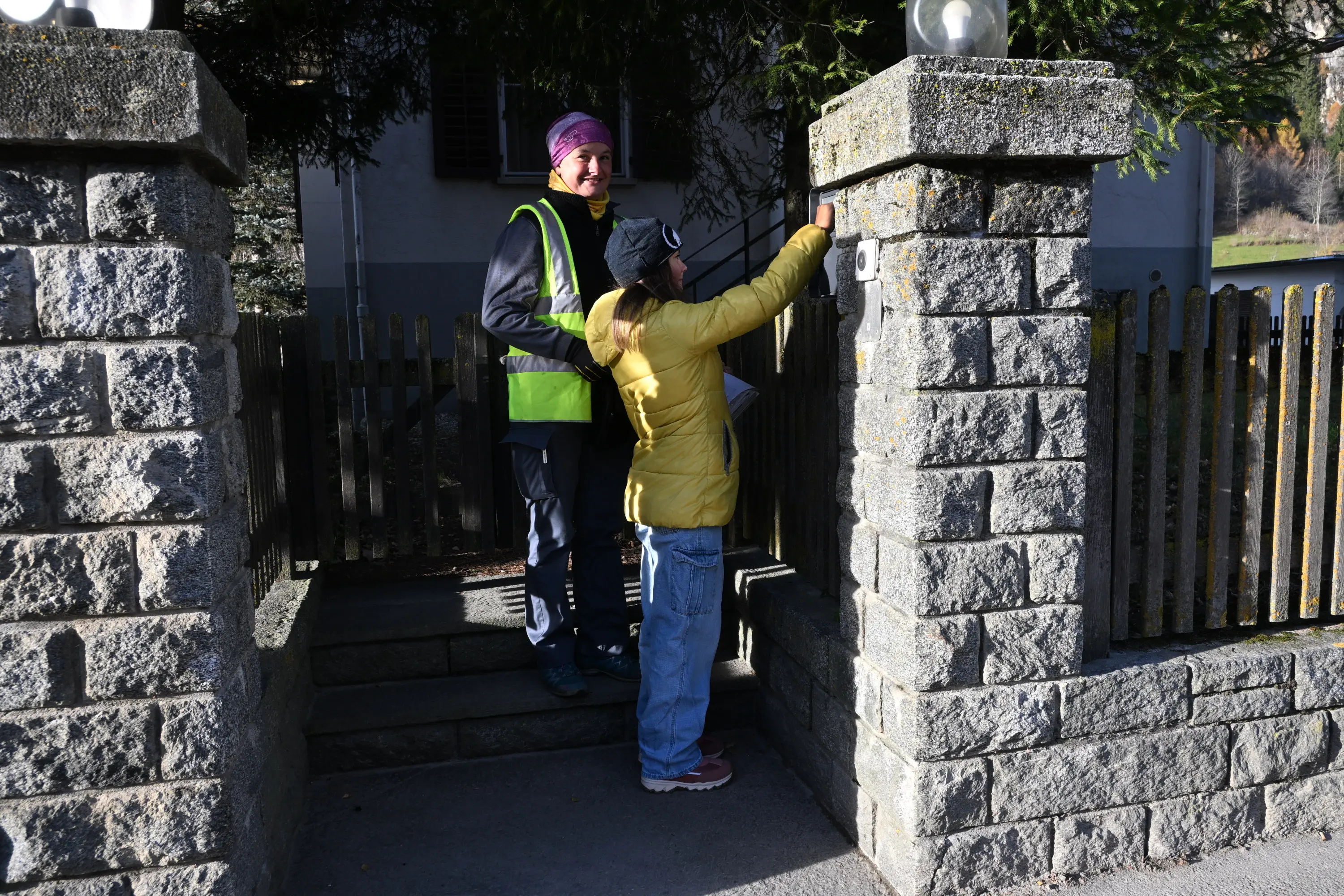 Due persone interagiscono davanti a un cancello: una indossa una giacca gialla e l'altra una giacca riflettente. La persona in giallo sta toccando il campanello, mentre l'altra sorride. Sullo sfondo si vede una casa e un