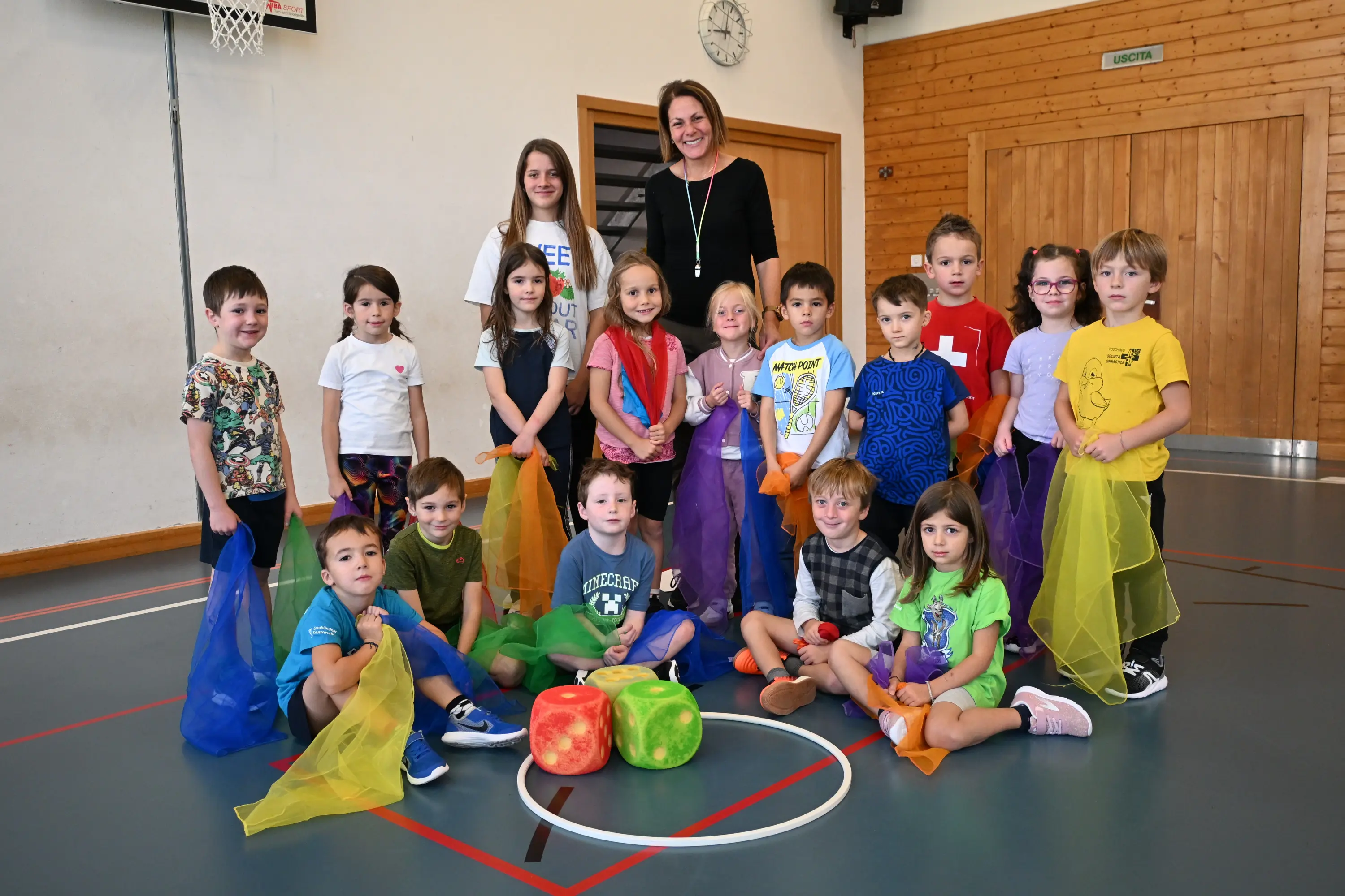 Gruppo di bambini sorridenti in palestra, con una donna adulta in piedi al centro. I bambini indossano foulard colorati e sono seduti o in piedi attorno a due grandi palloni colorati.
