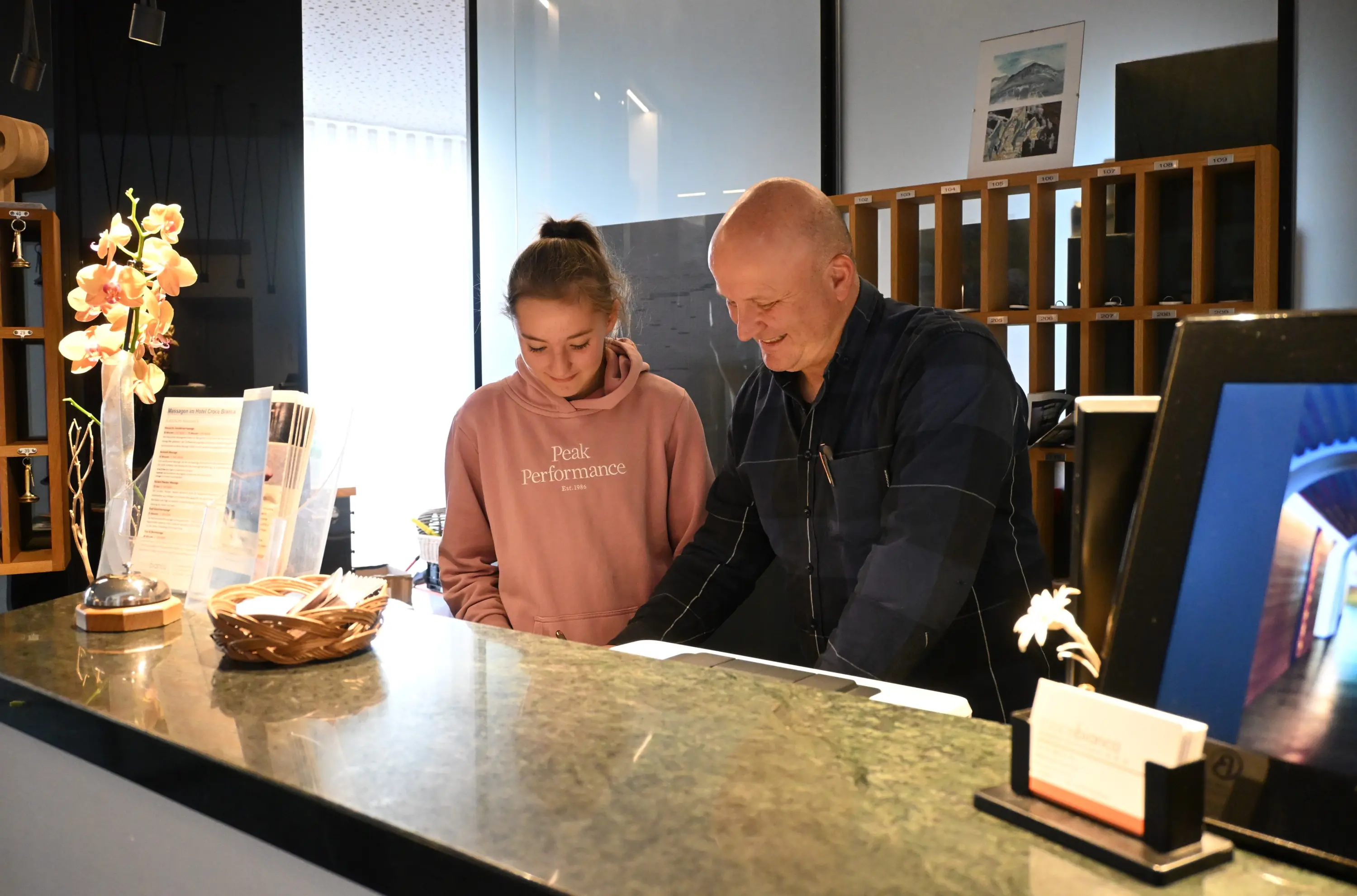 Due persone si trovano dietro un bancone di reception. Un uomo e una donna giovane stanno osservando un documento insieme, sorridendo. Il bancone è decorato con un vaso di fiori e ci sono materiali informativi in vista.