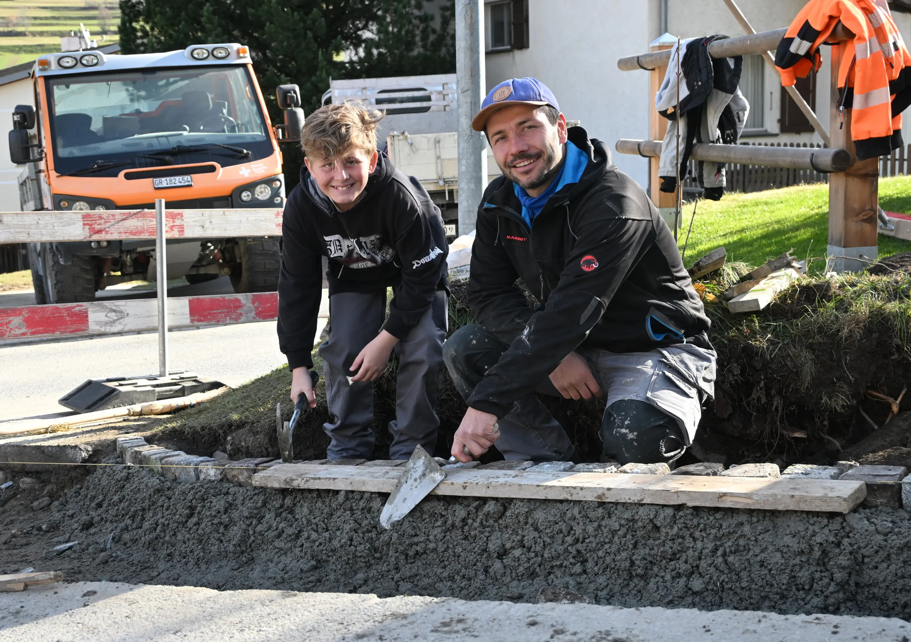 Due uomini sorridenti, uno adulto e uno giovane, stanno lavorando a un progetto di costruzione. Entrambi tengono delle cazzuole e sono seduti su una base di cemento. Sullo sfondo si intravede un camion arancione e delle barriere stradali.