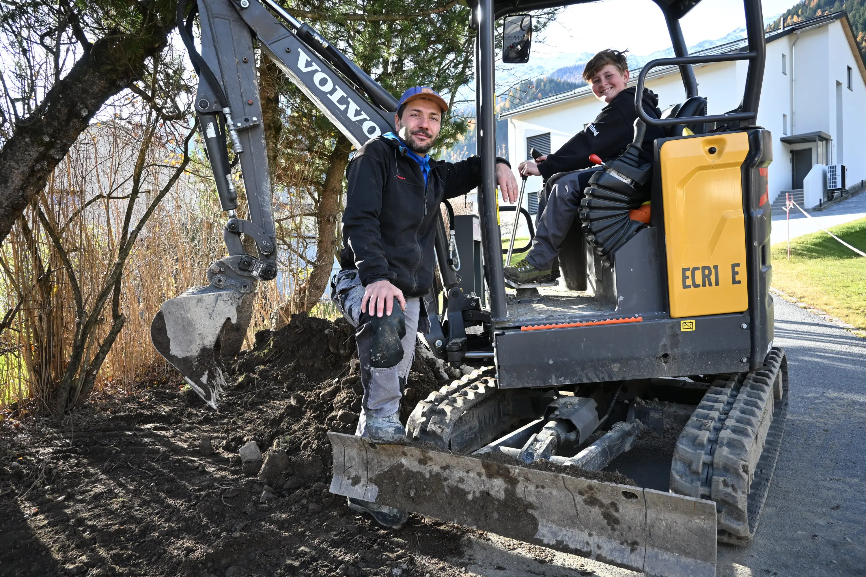 Due uomini sorridenti si trovano accanto a un escavatore giallo, uno è seduto in cabina e l'altro è in posizione accovacciata davanti alla macchina, sullo sfondo ci sono alberi e case.