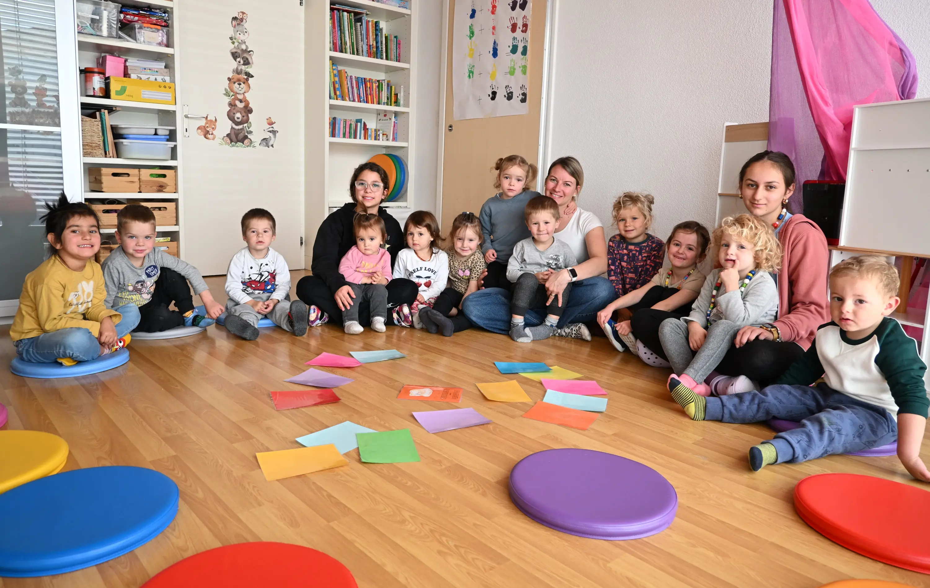 Un gruppo di bambini e adulti seduti in cerchio su un pavimento di legno, circondati da cuscini colorati. Alcuni bambini giocano con fogli colorati sparsi a terra, mentre gli adulti li osservano con attenzione. Sulla parete sono
