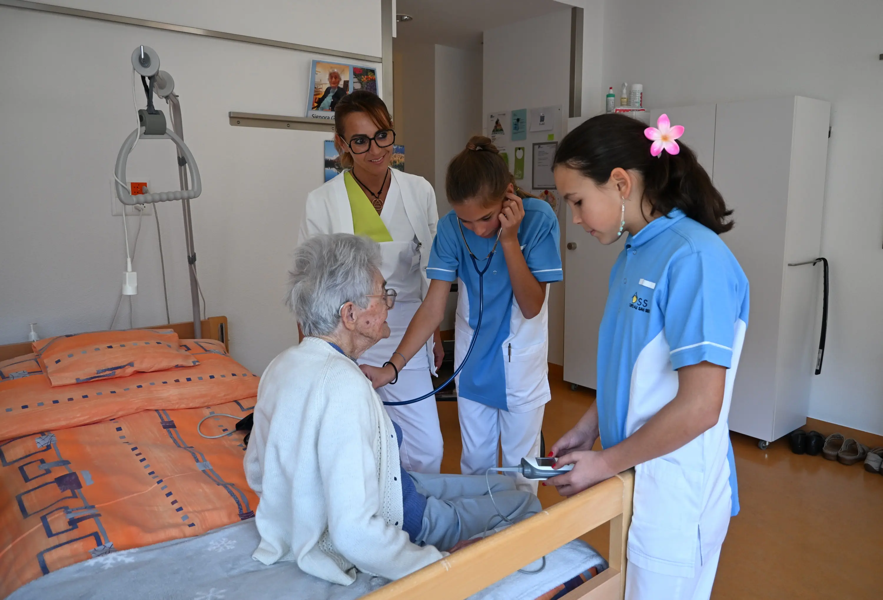 Un'anziana è seduta su un letto in una stanza, mentre due ragazze in uniforme medica le controllano la pressione sanguigna. Una professionista sanitaria osserva e ascolta attentamente.
