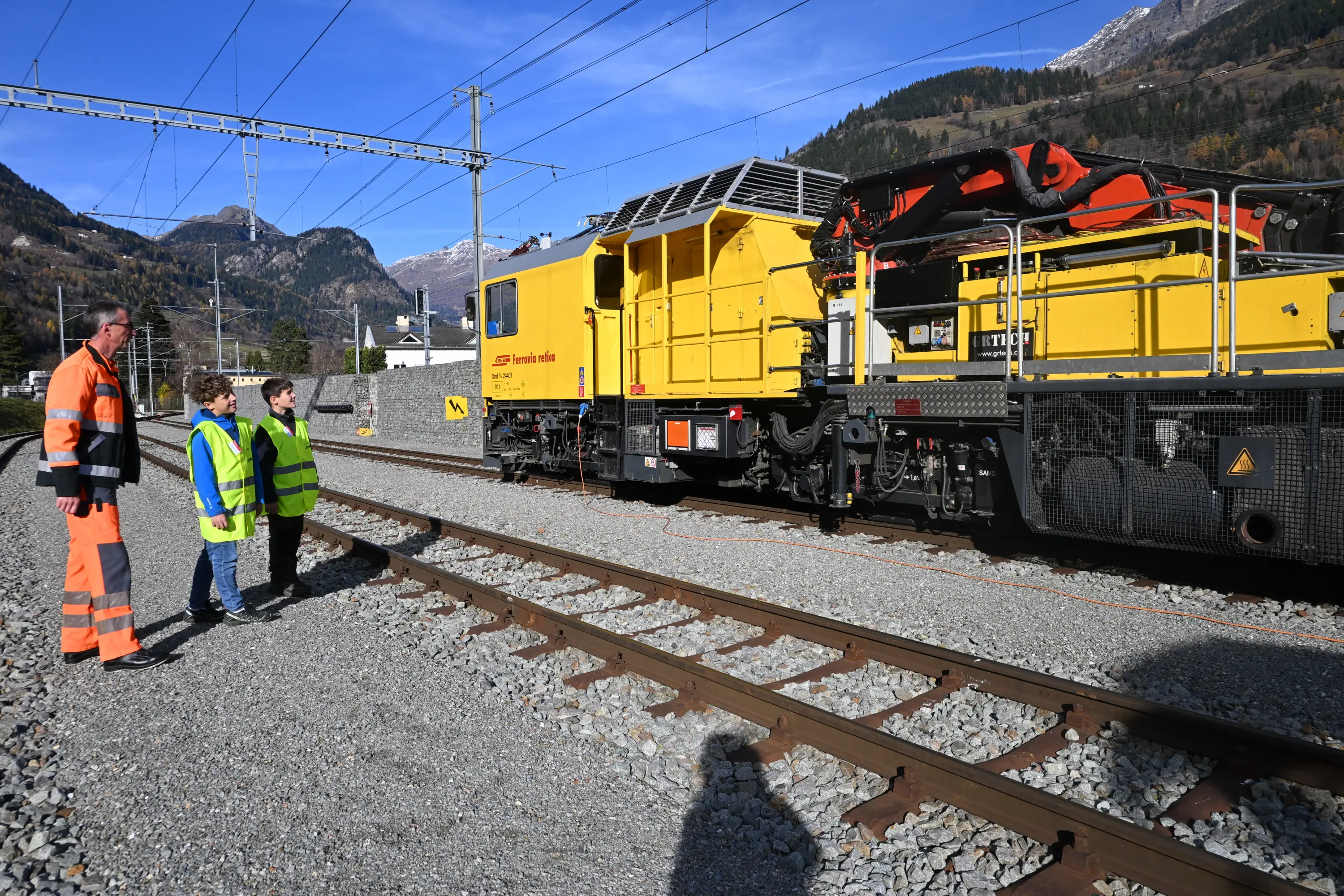 Tre persone in abbigliamento fluorescente osservano un treno giallo su un binario, con montagne sullo sfondo. Un operatore è vicino a un macchinario montato sul treno.