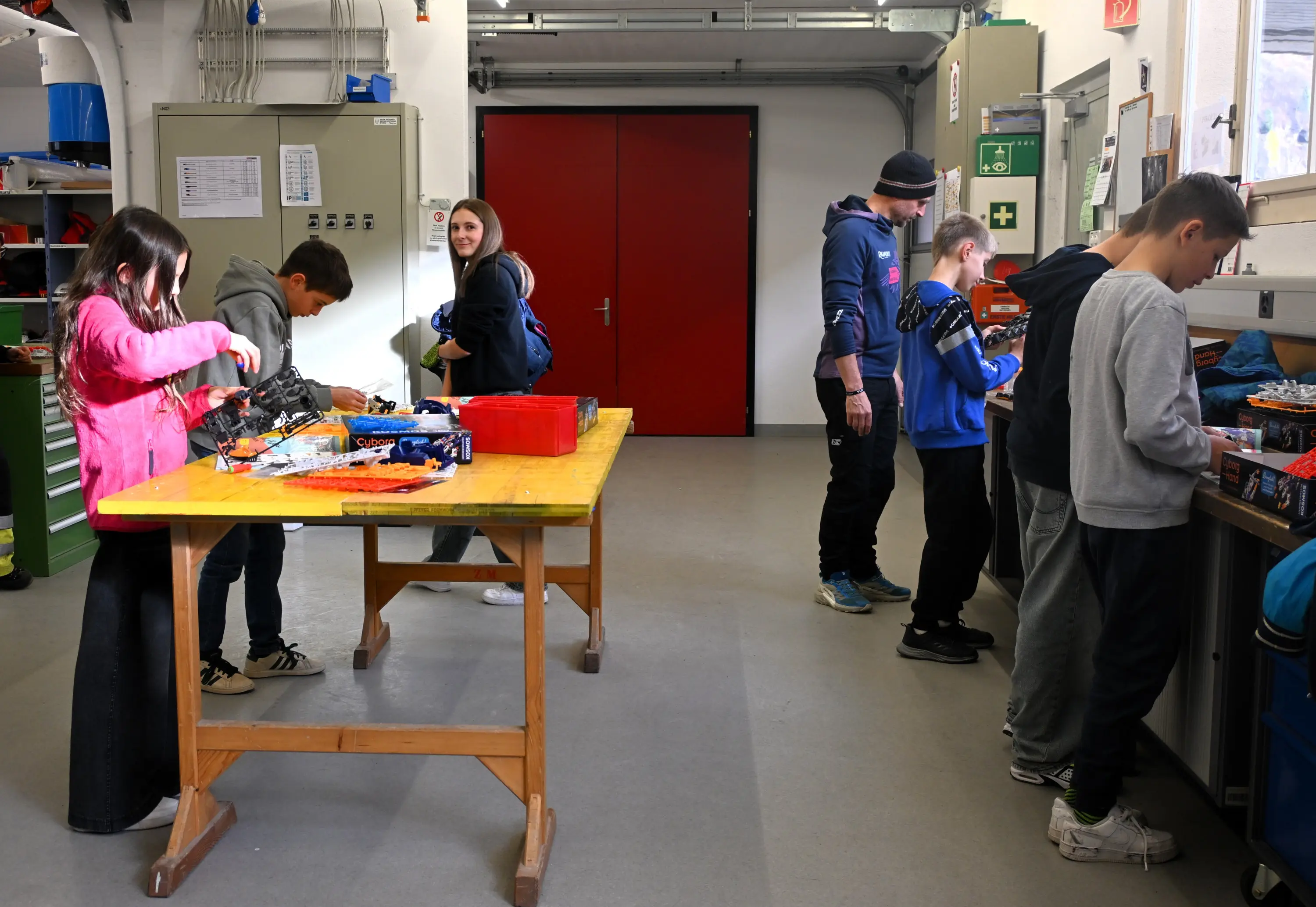 Un gruppo di ragazzi lavora in un laboratorio. Alcuni sono impegnati con attrezzi su un tavolo centrale, mentre altri si trovano a una bancarella laterale, tutti concentrati sulle loro attività. Sullo sfondo ci sono armadietti e attrezzature da