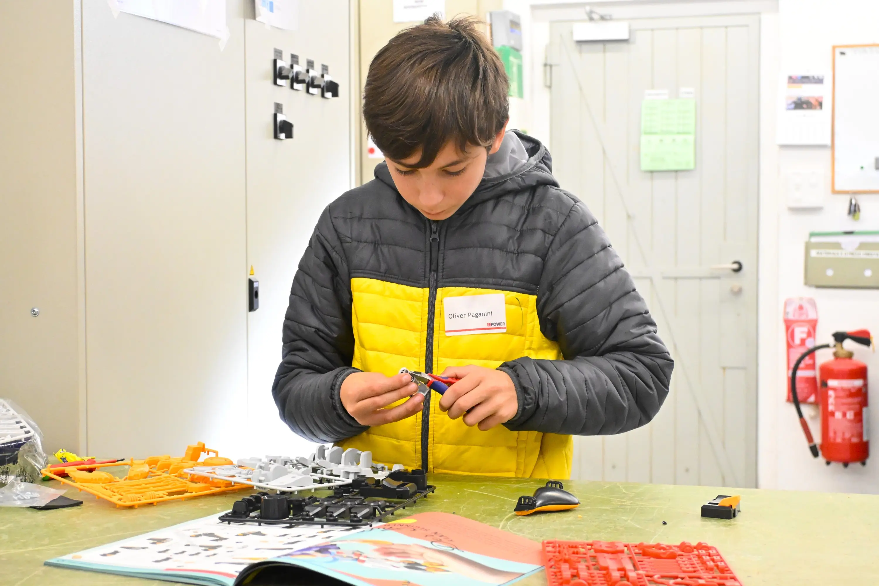 Un ragazzo con un giubbotto giallo e nero sta assemblando un modello con pezzi colorati su un tavolo, mentre ha un libro aperto davanti a sé.