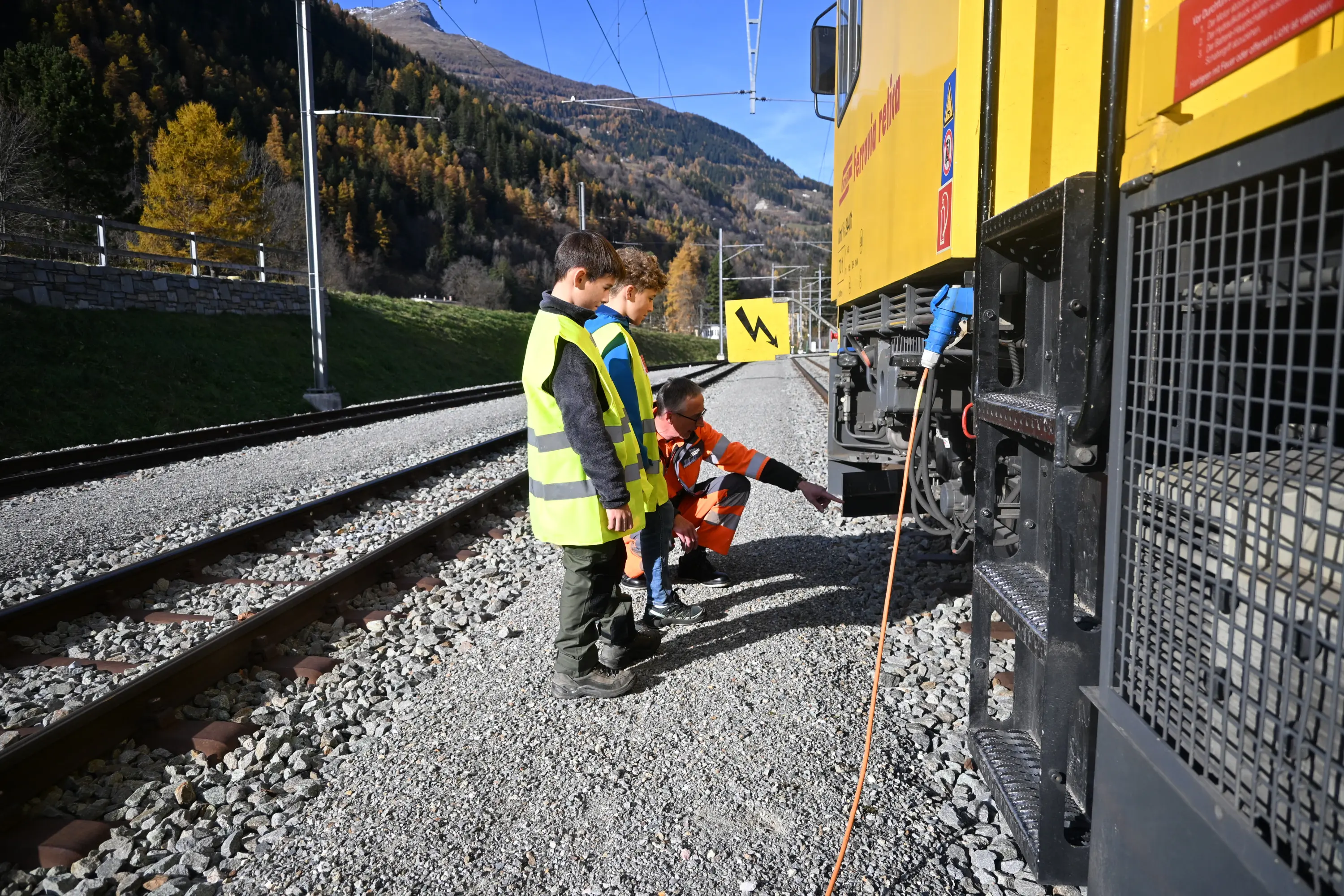 Due bambini vestiti con giubbotti riflettenti osservano un adulto in tuta da lavoro che punta verso il lato di un treno giallo. Sullo sfondo si vedono alberi con foglie autunnali e una linea ferroviaria.