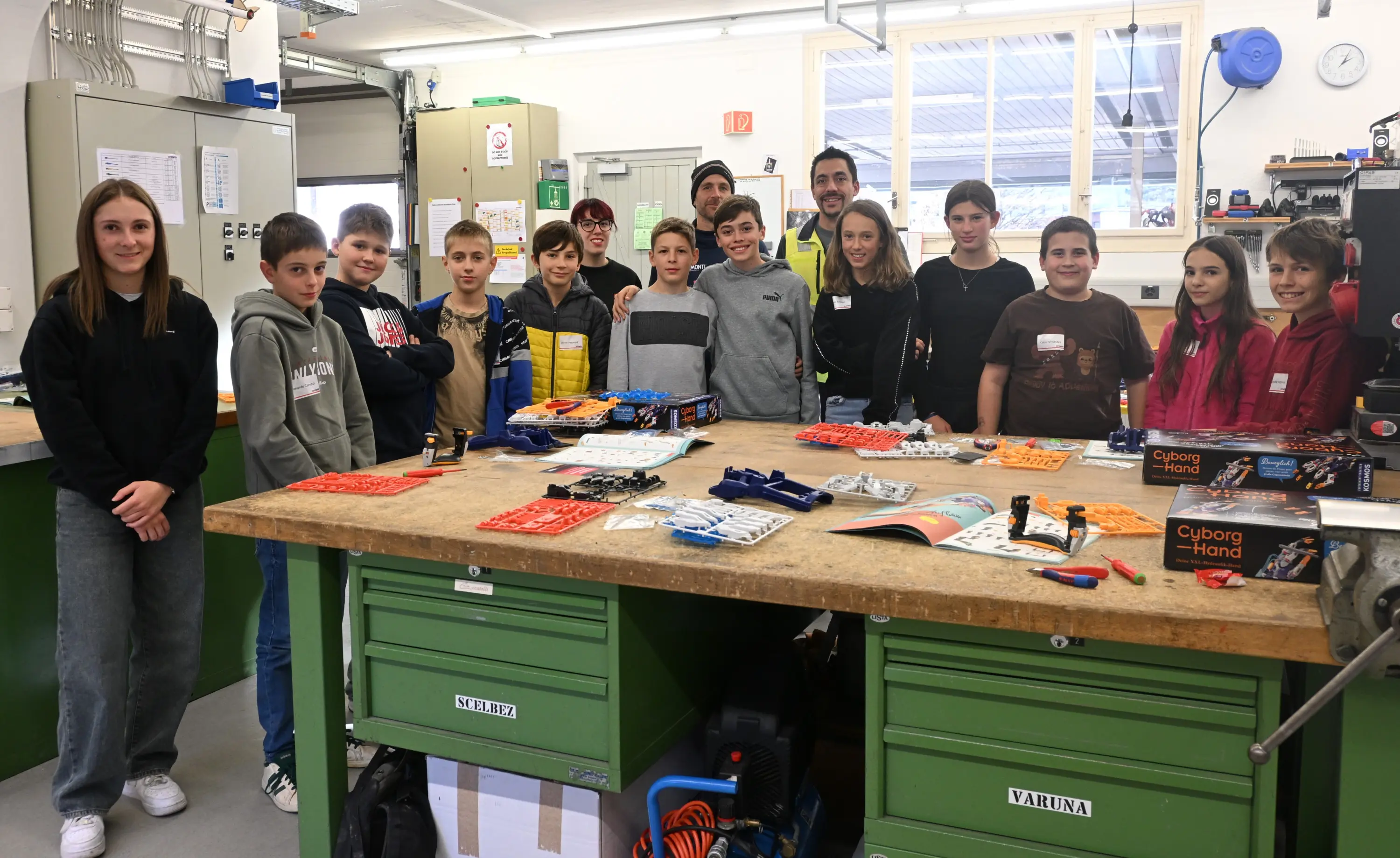 Gruppo di giovani studenti e un adulto in un laboratorio, sorridendo di fronte a un tavolo con attrezzature e materiali didattici. Sullo sfondo si vedono attrezzature e attrezzi vari.