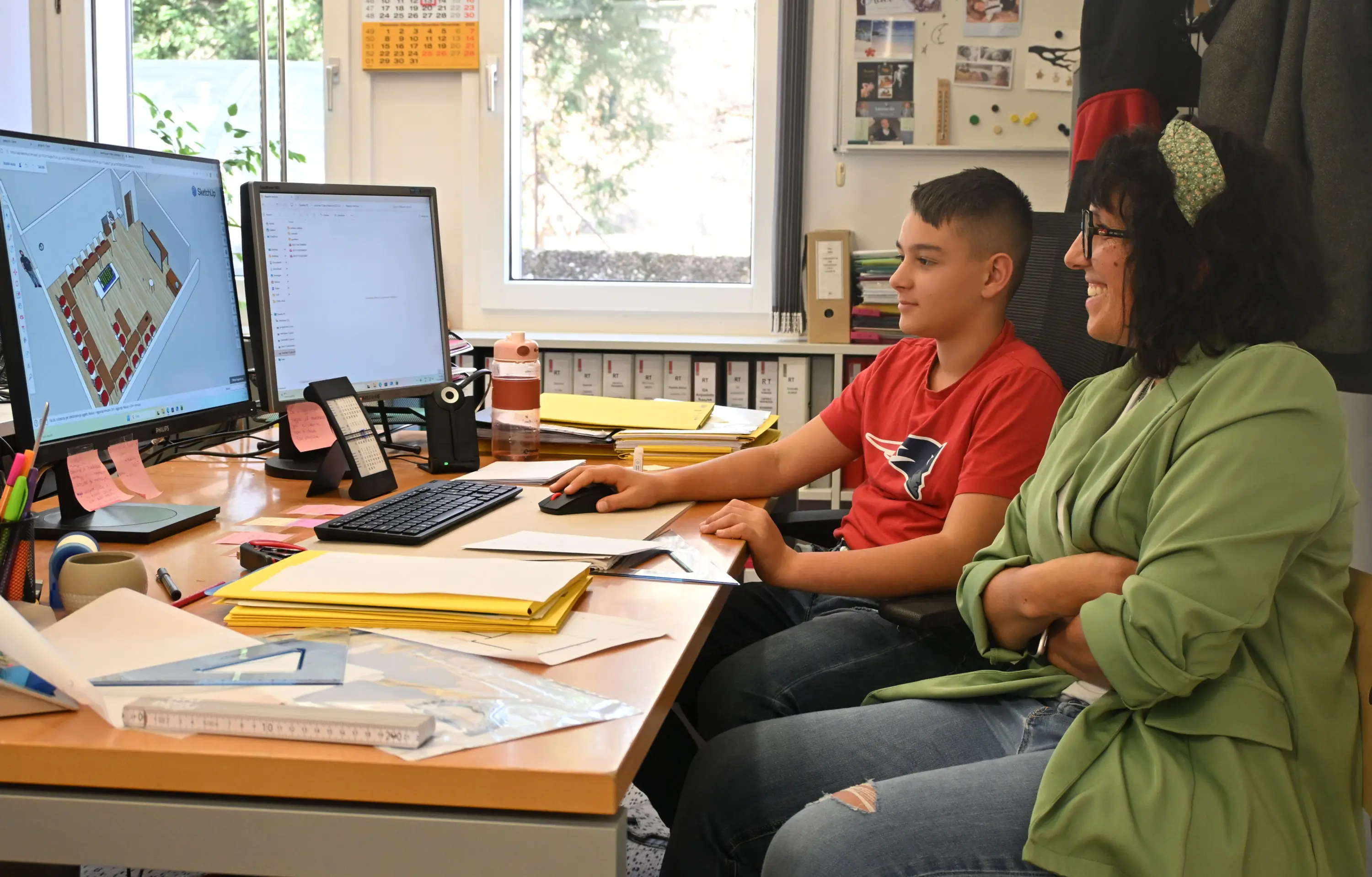 Un ragazzo e una donna sorridente seduti a una scrivania, entrambi osservano uno schermo di computer. Sullo schermo è visibile un progetto di design d'interni. La scrivania è disordinata con fogli, cancelleria e una bott