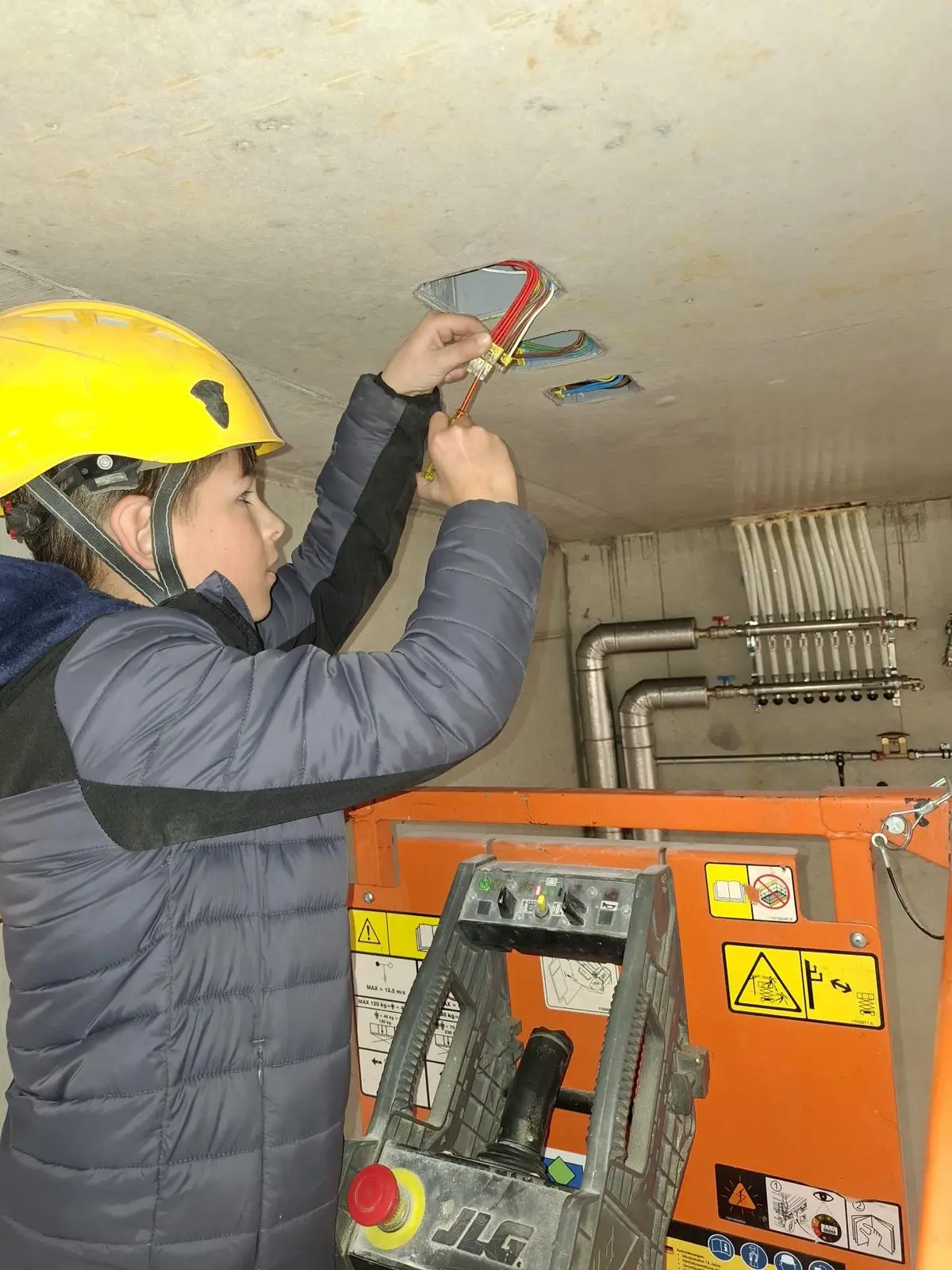Un bambino con elmetto giallo sta lavorando su un edificio, mentre utilizza strumenti per collegare tubi al soffitto in una stanza con tubature a vista.