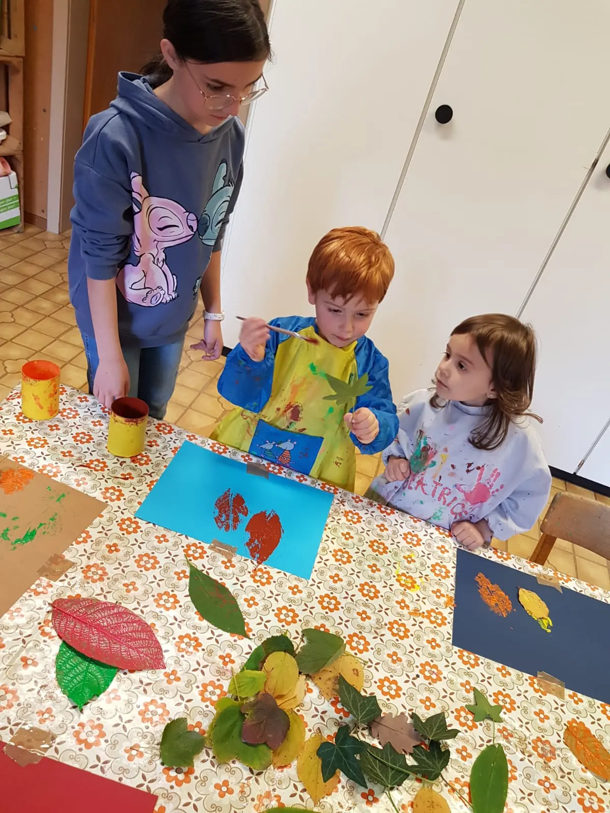 Due bambini, uno con i capelli rossi e un grembiule, stanno dipingendo foglie su un foglio blu. Una ragazza più grande li osserva. Sul tavolo ci sono colori e foglie.