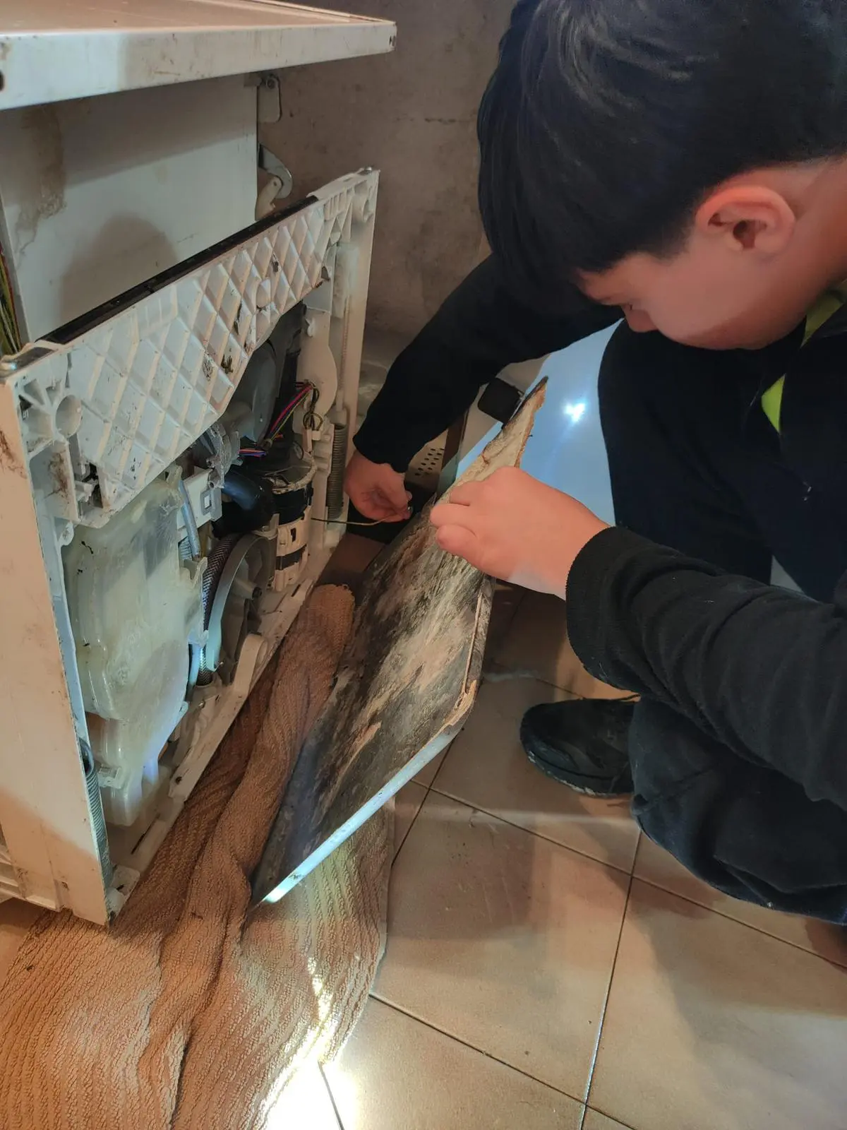 Un ragazzo sta lavorando su un elettrodomestico, rimuovendo un pannello laterale mentre tiene in mano un attrezzo. Sul pavimento è presente una coperta.