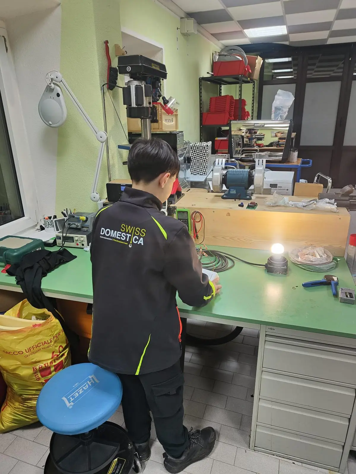 Un ragazzo lavora a un banco da lavoro, con attrezzi e materiali disposti ordinatamente. Sullo sfondo si vedono scaffali con attrezzature e un'illuminazione adeguata.