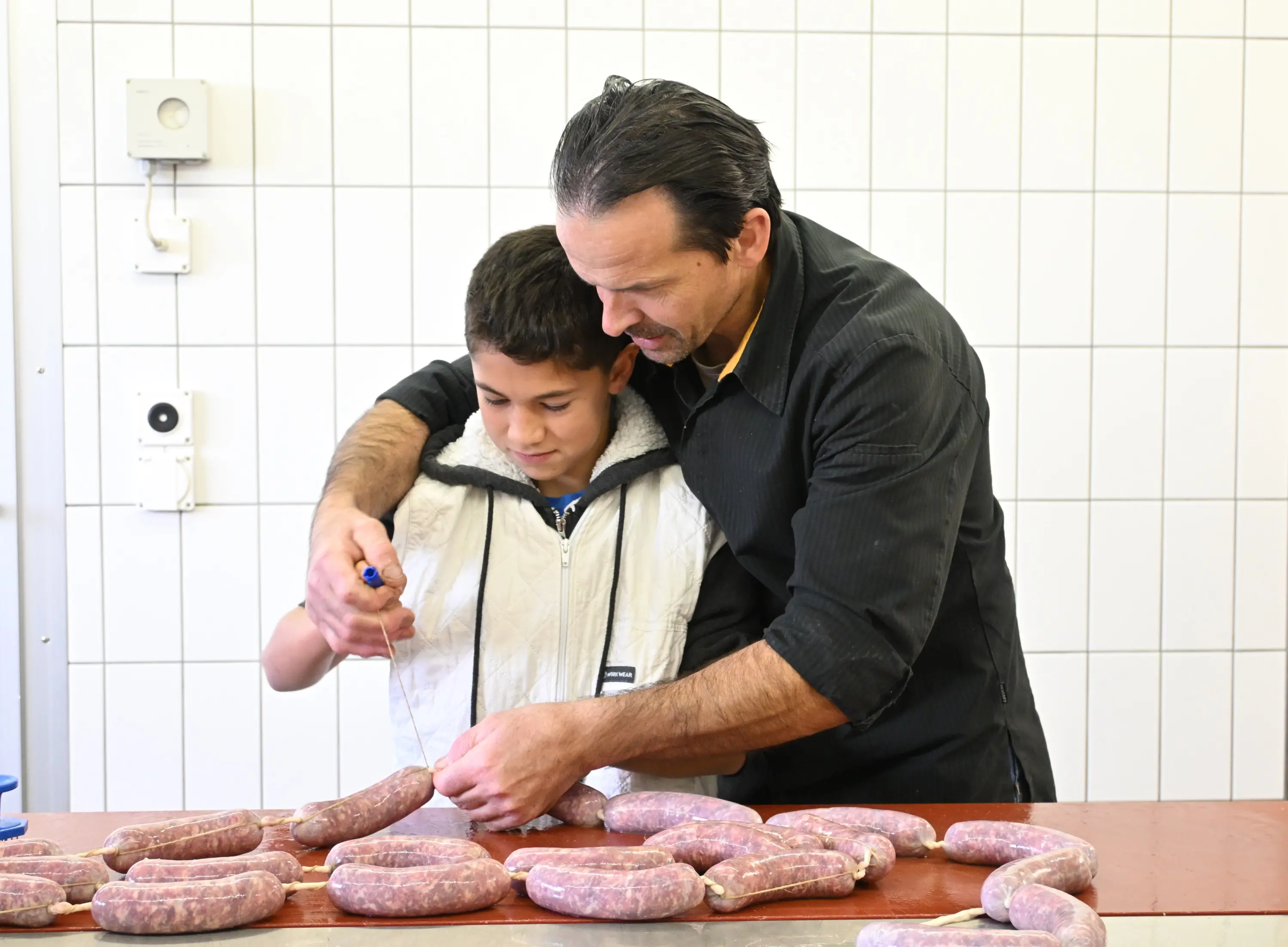 Un uomo e un ragazzo stanno preparando salsicce su un tavolo di lavoro. L'uomo tiene la mano del ragazzo mentre entrambi scrivono su una salsiccia. Intorno a loro ci sono diverse salsicce già formate.