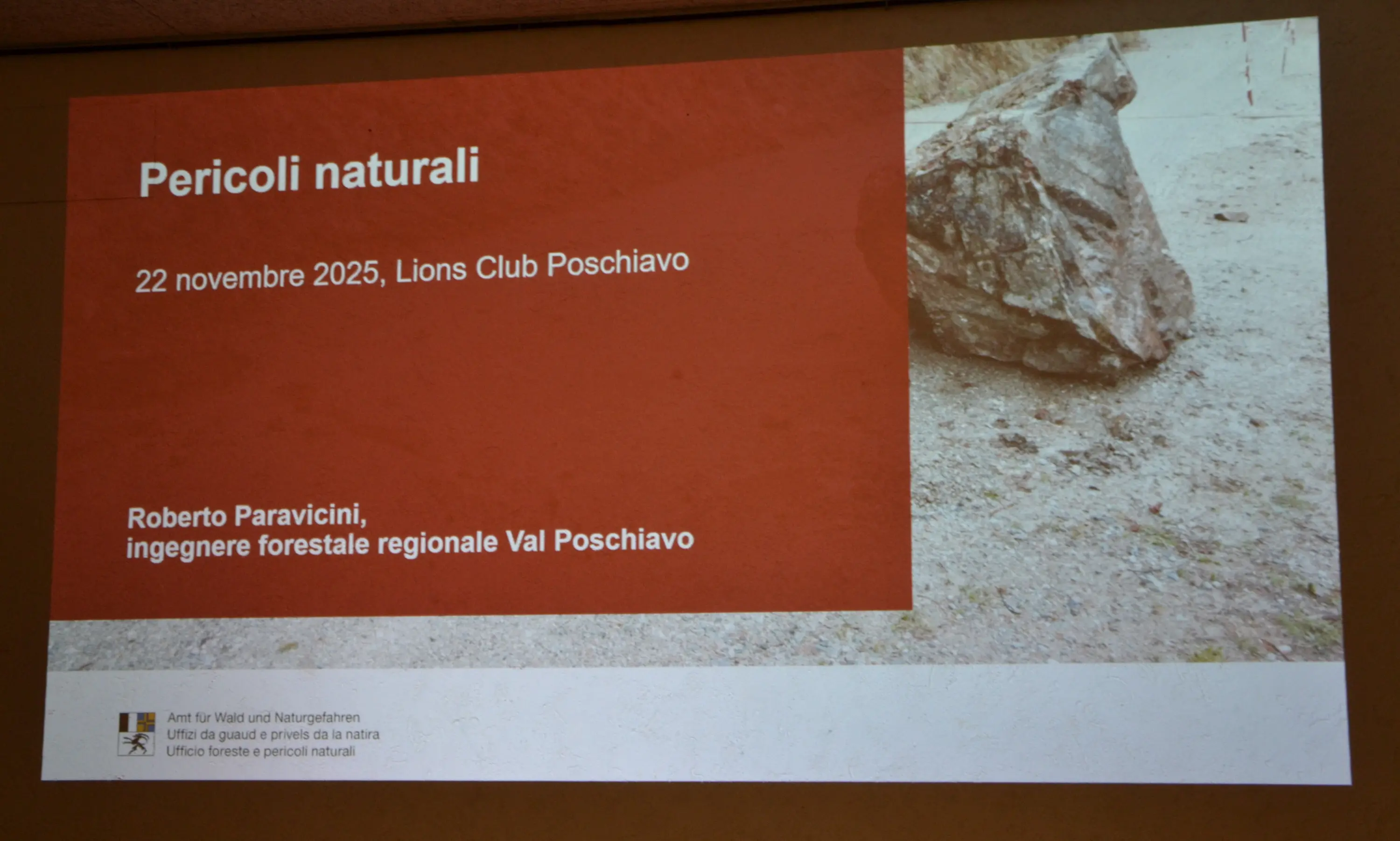 Presentazione intitolata "Pericoli naturali" con data e luogo. Include il nome del relatore, Roberto Paravicini, ingegnere forestale regionale Val Poschiavo. Sfondo con un sasso e un terreno naturale.