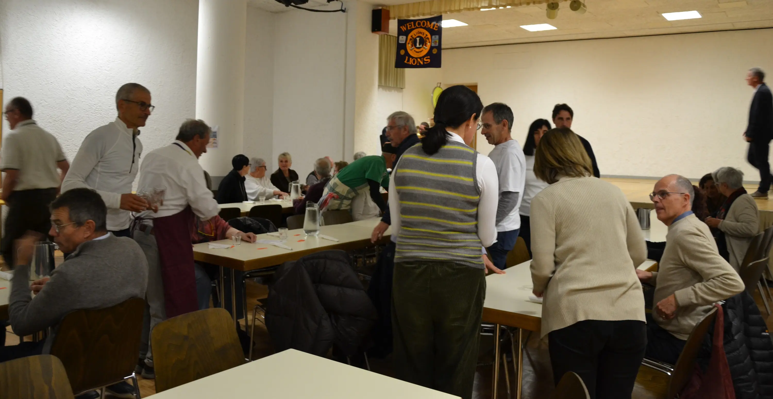Un gruppo di persone adulte si riunisce in una sala, alcune conversano mentre altre sistemano tavoli e sedie. Si nota un'atmosfera conviviale, con una parete decorata e un palco sullo sfondo.