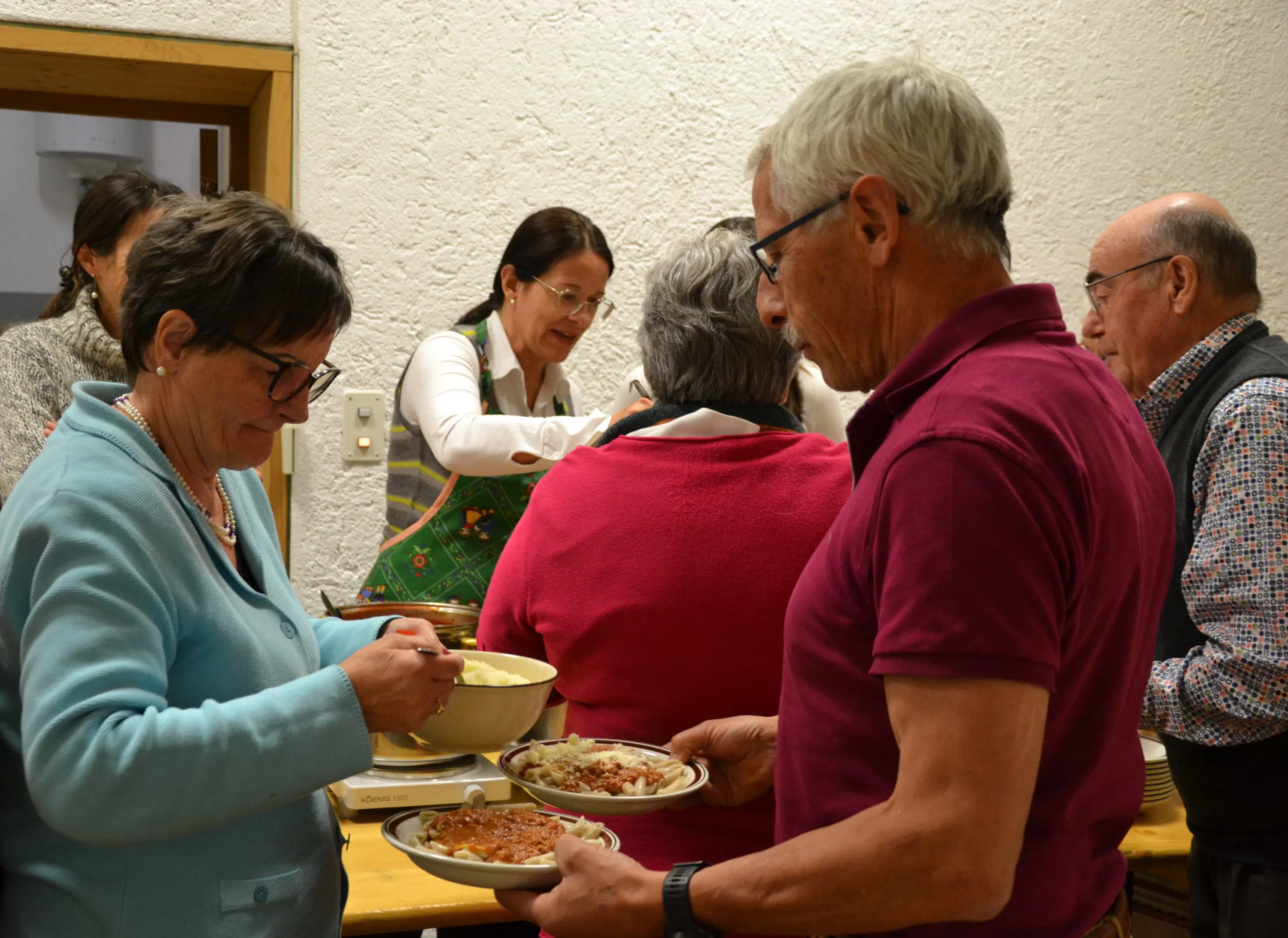 Un gruppo di persone si è riunito per un pasto. Due persone in primo piano stanno servendo il cibo nei piatti, mentre altre sono in attesa dietro di loro. Una donna indossa un grembiule e sta preparando i piatti.
