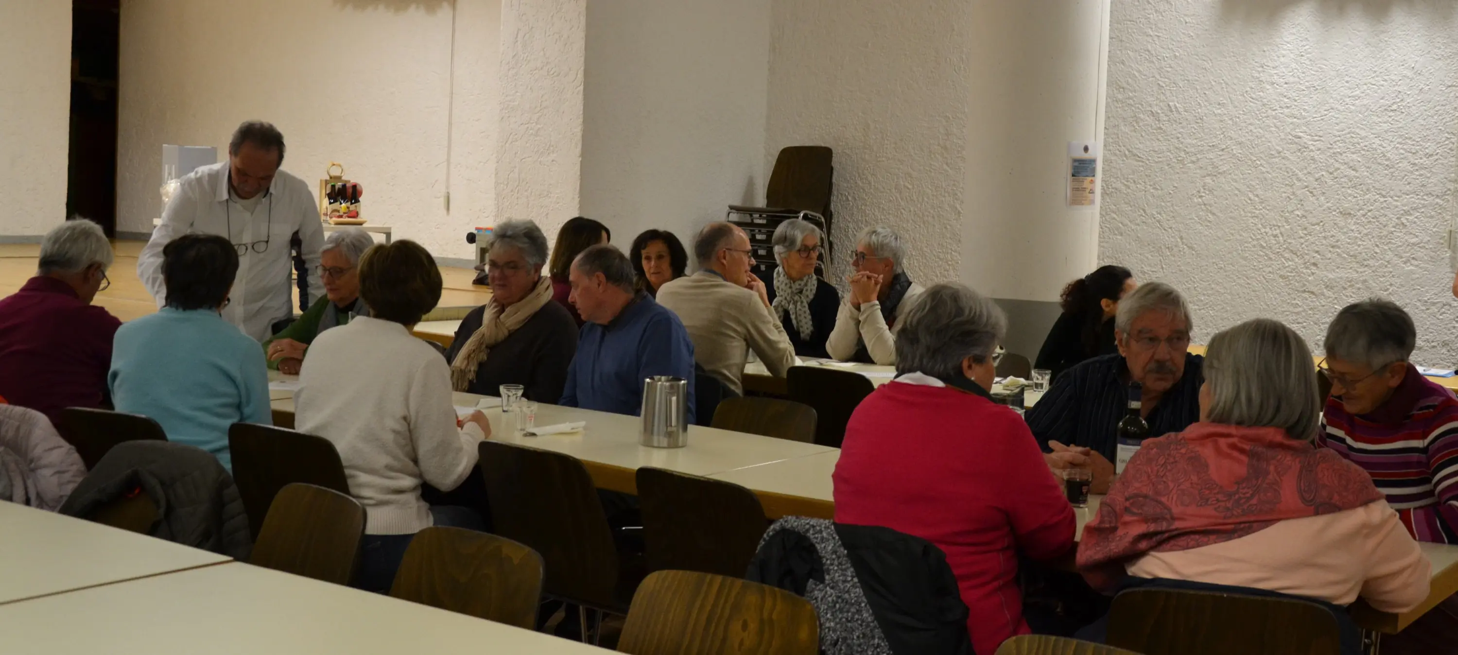 Un gruppo di persone anziane si riunisce in una sala, sedute attorno a tavoli. Alcuni conversano, mentre altri bevono e mangiano. L'atmosfera è conviviale e calorosa.