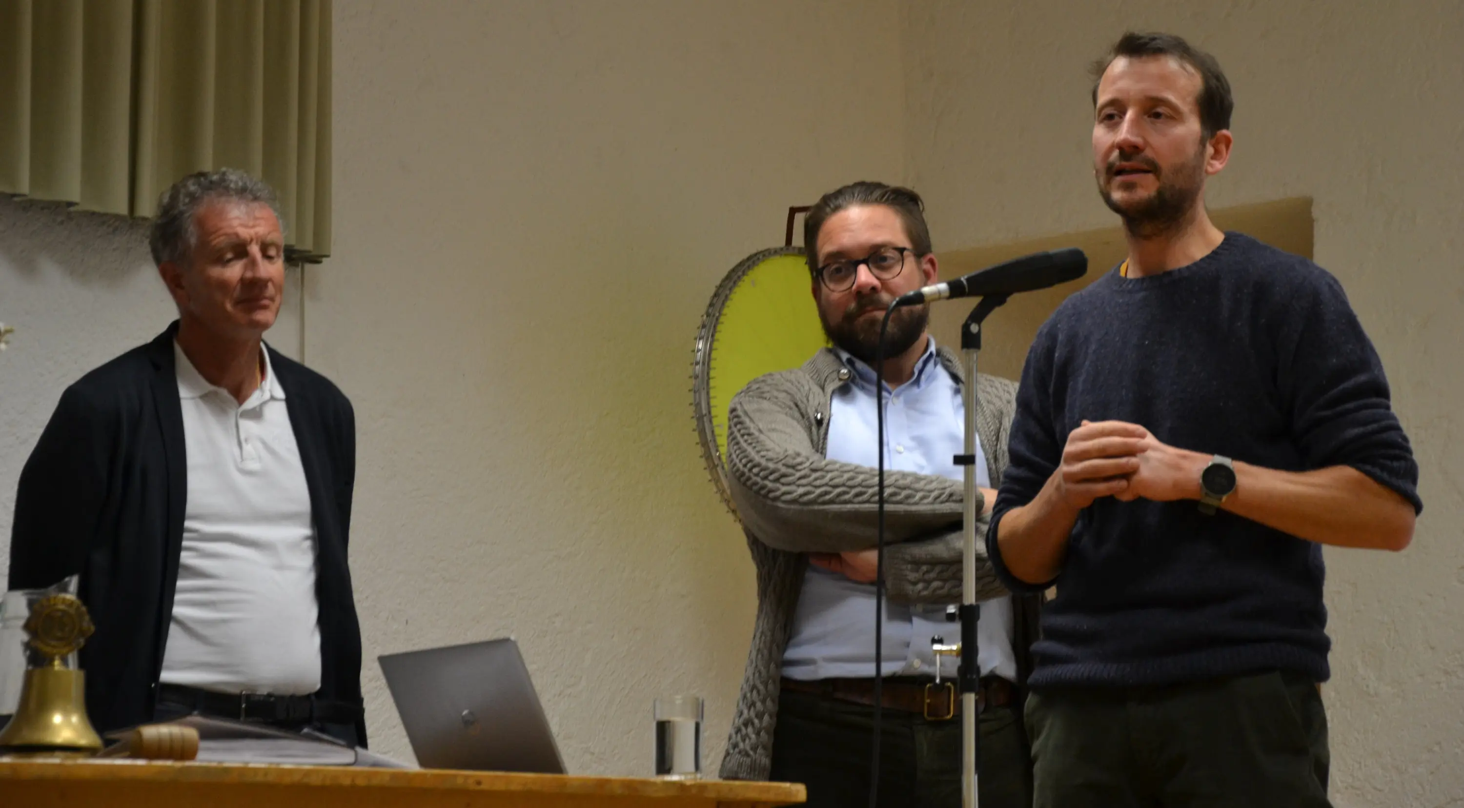 Tre uomini in una sala, con uno in piedi davanti a un microfono che parla. Gli altri due ascoltano; uno indossa un maglione e gli occhiali, l'altro ha le mani in tasca. Un computer portatile è visibile su un tavolo.