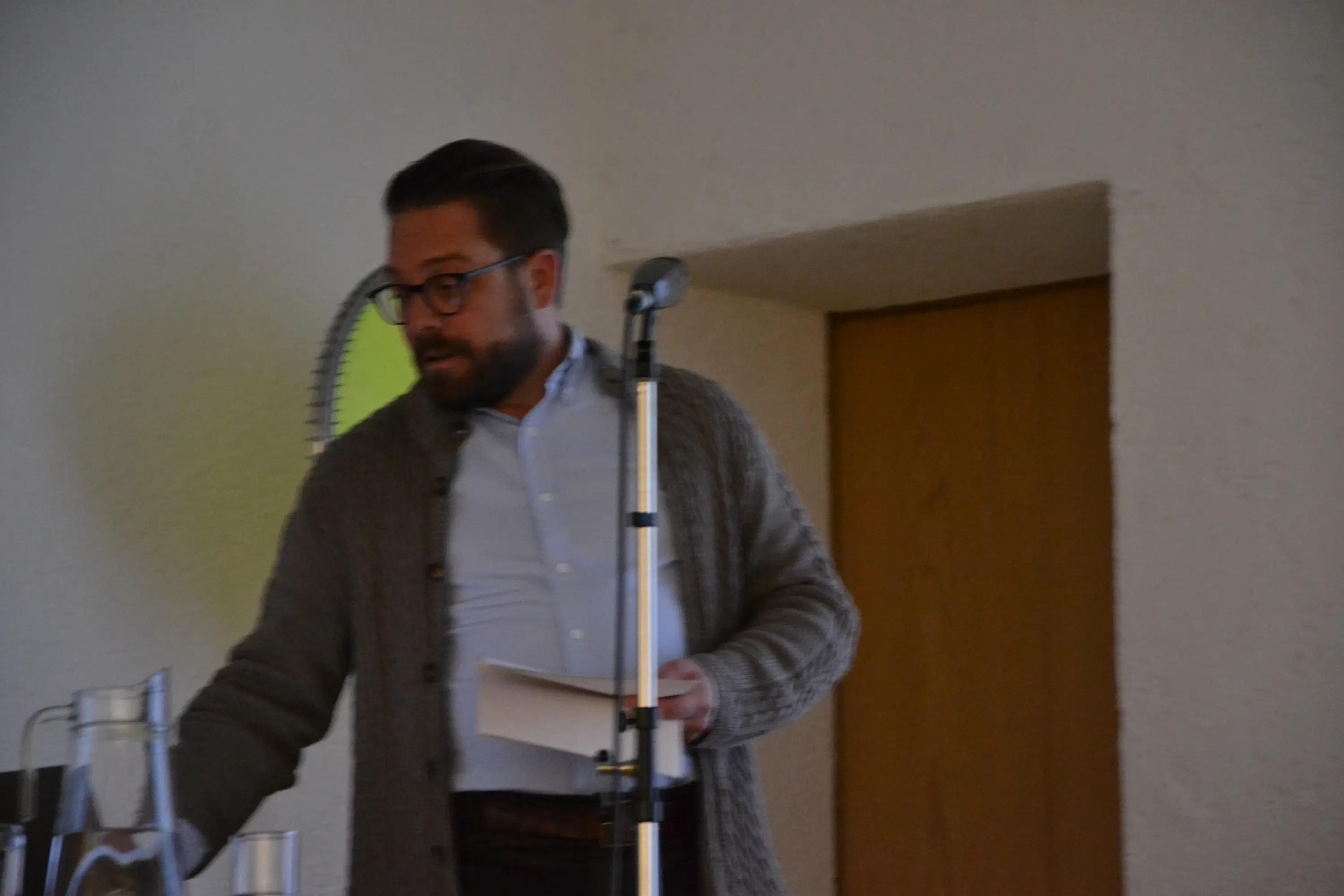 Uomo con occhiali e maglione grigio si avvicina a un tavolo con bottiglie d'acqua e un microfono. Sullo sfondo si intravede una porta.