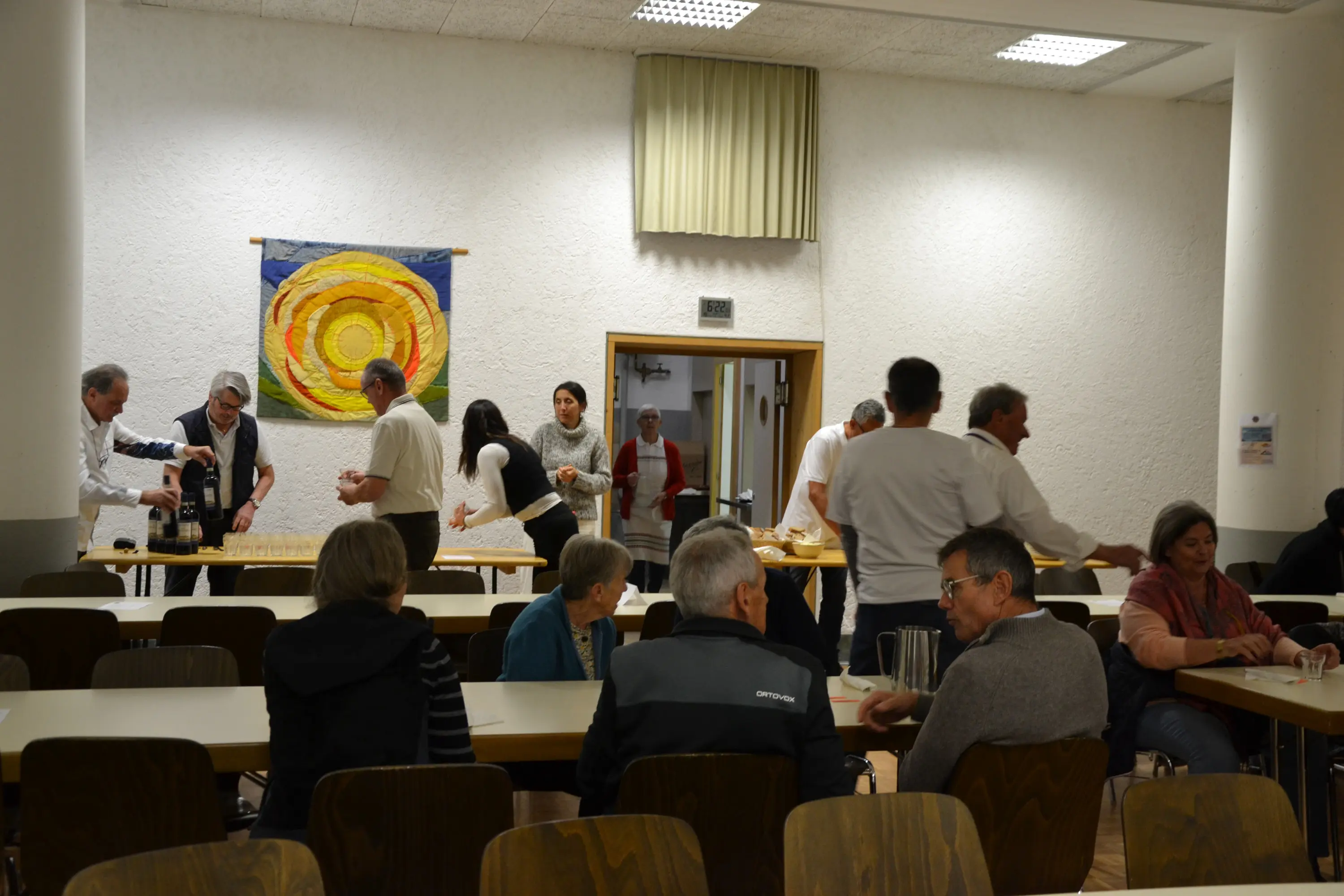 Una sala con diverse persone che interagiscono e preparano tavoli. In primo piano, alcuni gruppi di persone siedono ai tavoli, mentre sullo sfondo si vedono persone in movimento e un grande arazzo colorato.