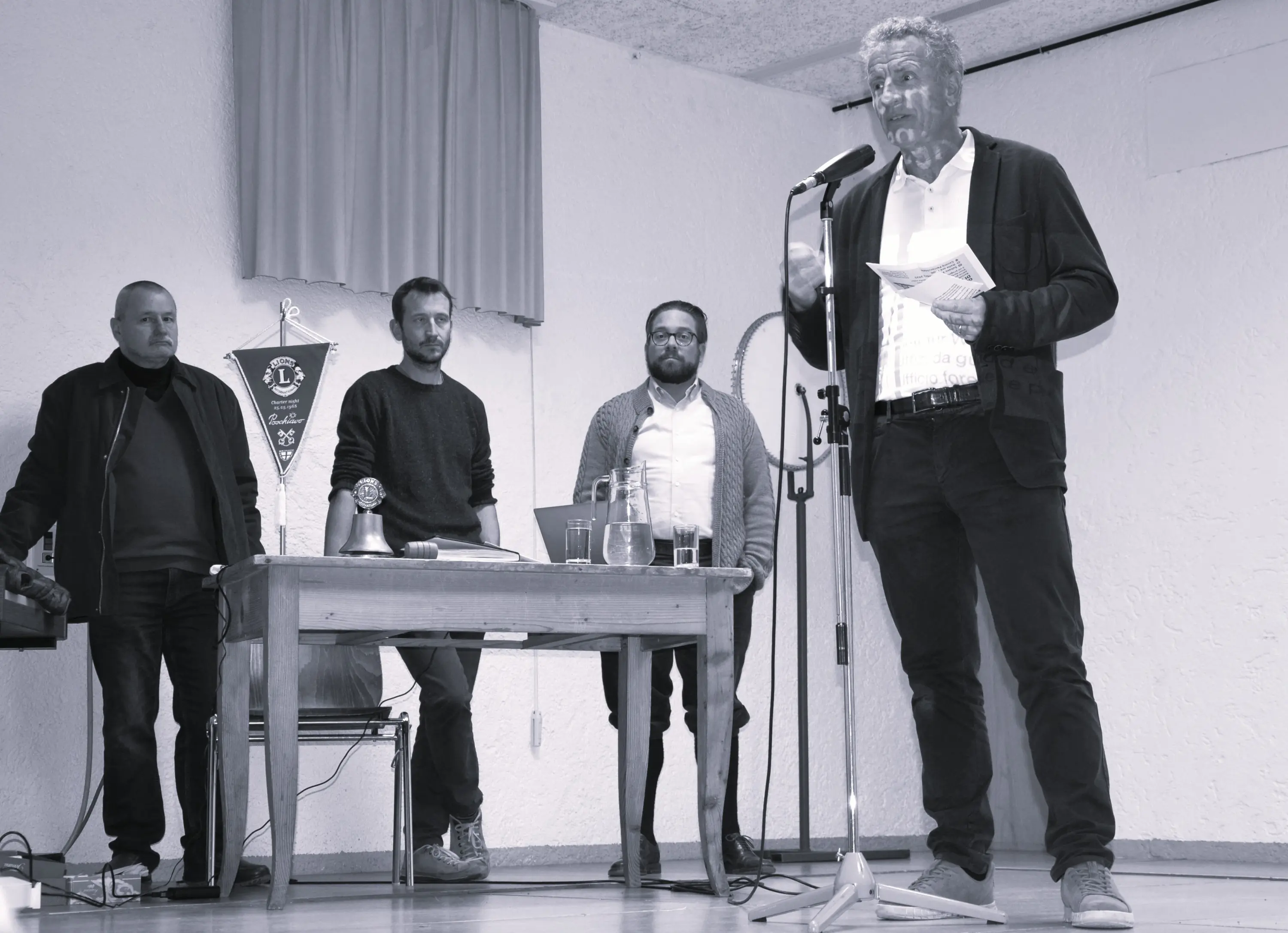 Un gruppo di quattro uomini si trova in una sala; uno di loro, in piedi, parla al microfono. Gli altri tre ascoltano attentamente, accanto a un tavolo con bicchieri e oggetti vari. La scena è in bianco e nero.