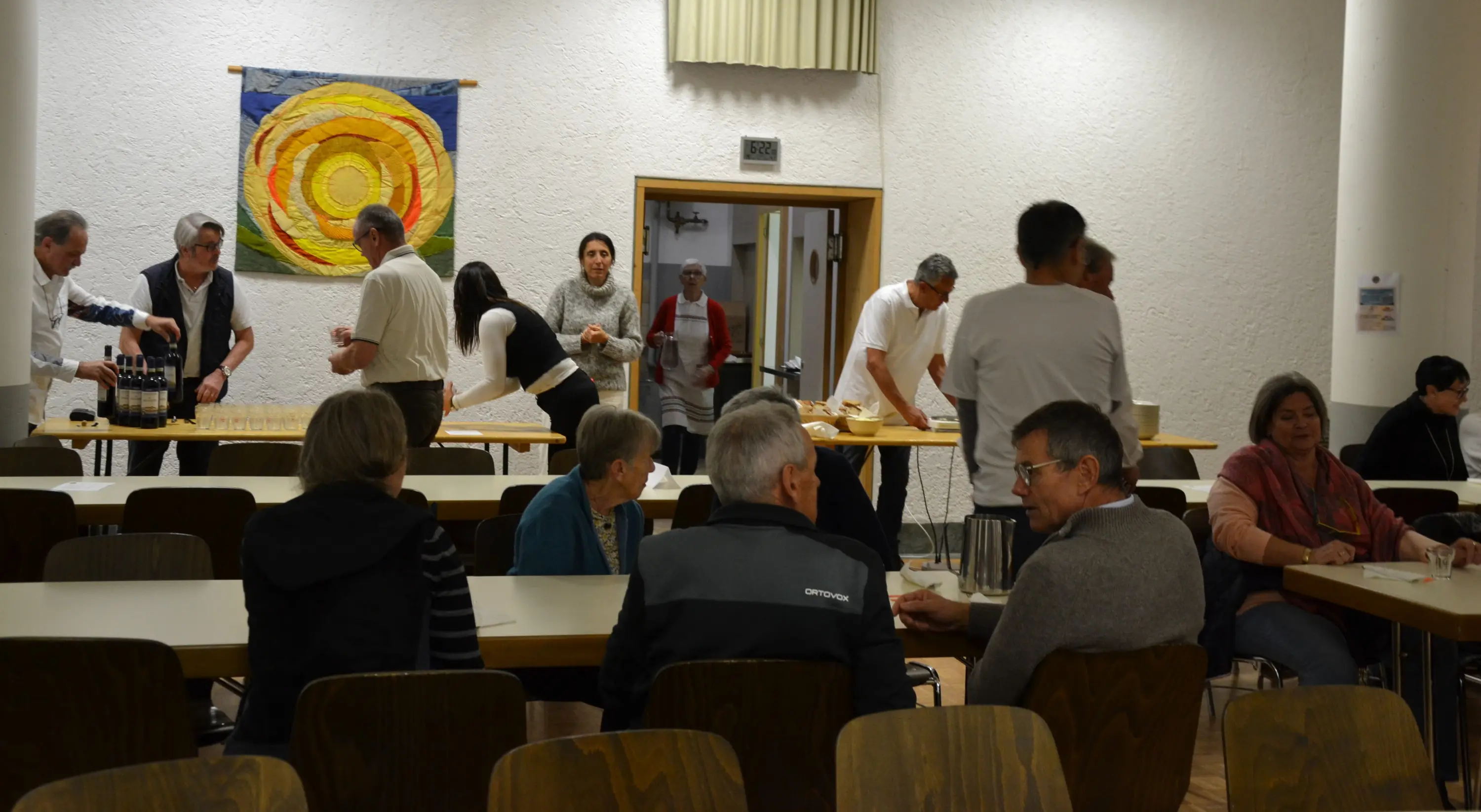 Un gruppo di persone in un ambiente di sala, alcune stanno preparando cibo e bevande, mentre altre sono sedute ai tavoli in conversazione. Un arazzo con un motivo circolare è appeso alla parete.