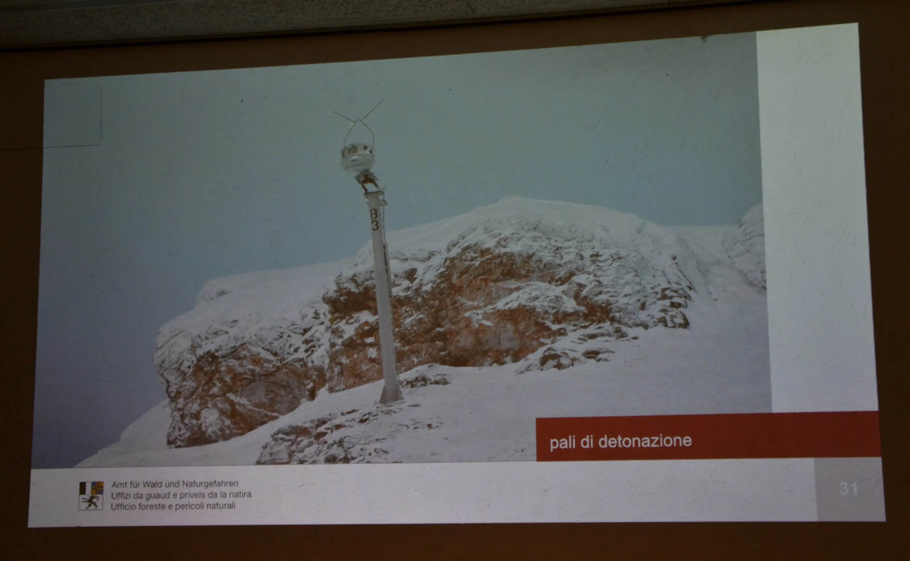 Struttura di detonazione situata su una montagna innevata, con un albero di sostegno e un mulino a vento. Atmosfera invernale con cielo nuvoloso.