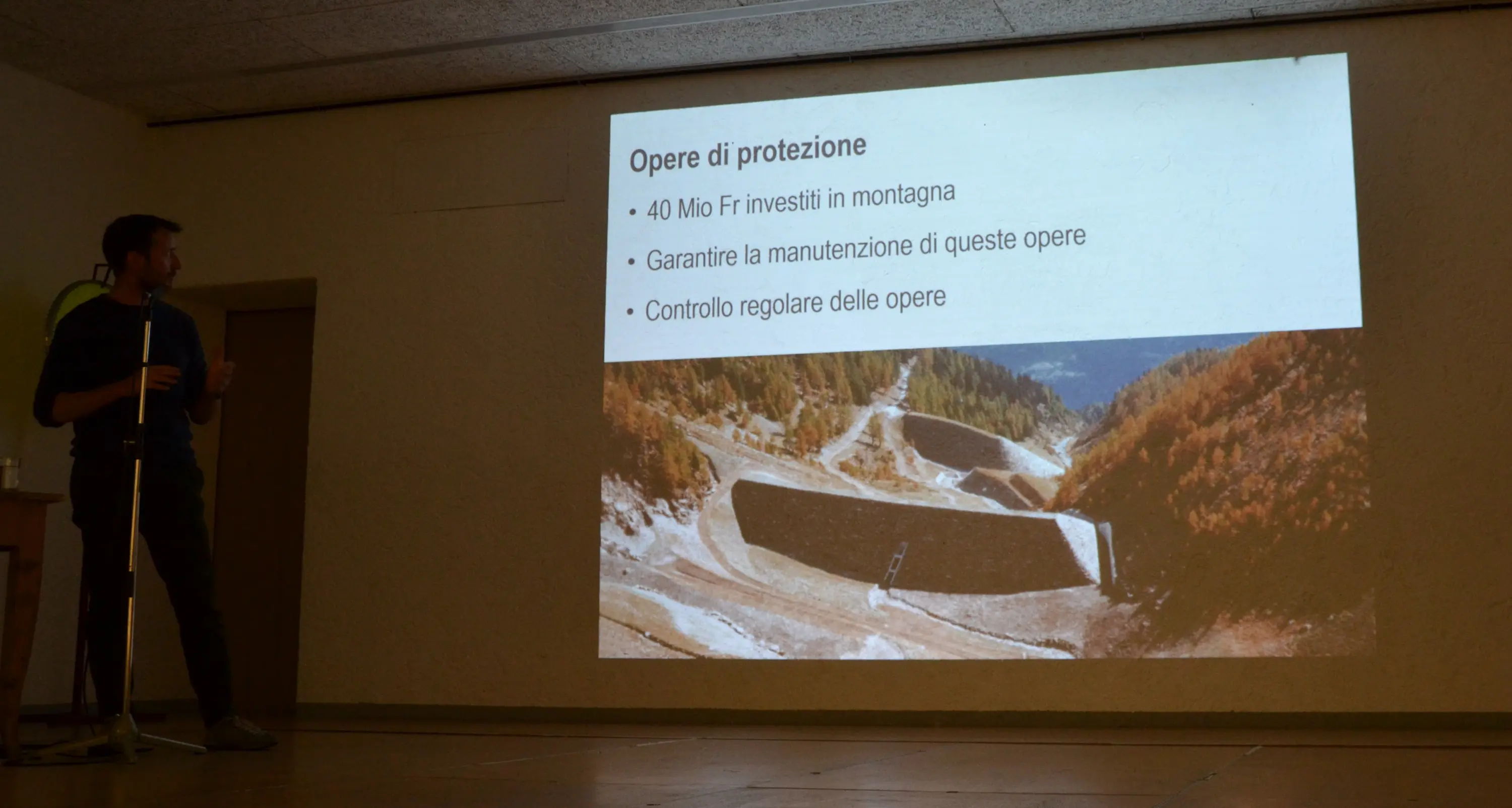 Un uomo presenta una diapositiva su opere di protezione, con un'immagine di una struttura montana. La diapositiva evidenzia investimenti, manutenzione e controllo regolare delle opere.