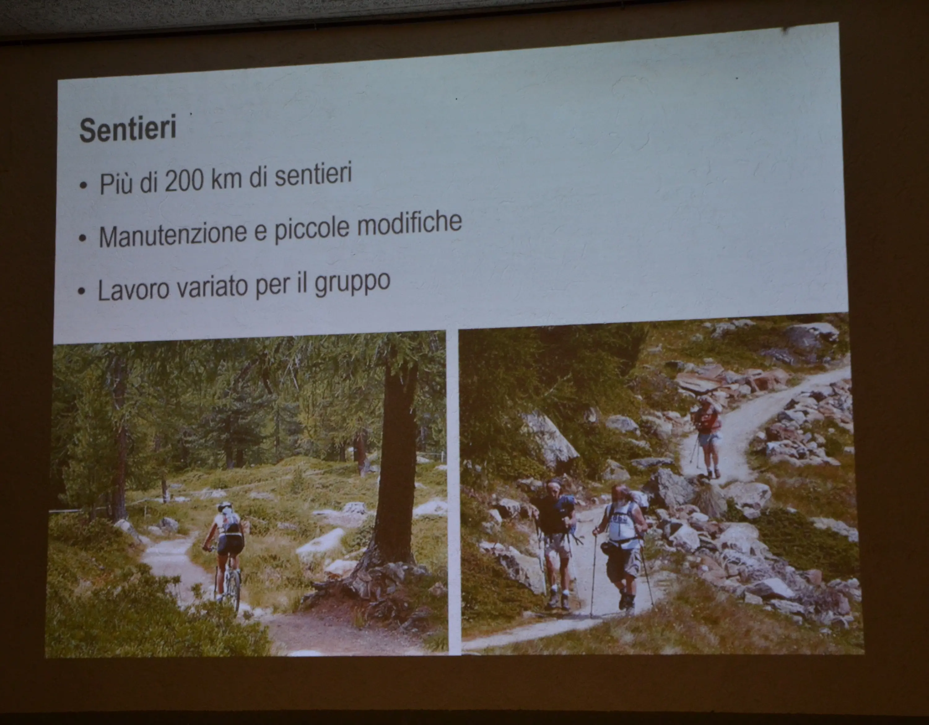 Due immagini su uno schermo che mostrano sentieri escursionistici. A sinistra, una persona in bicicletta su un sentiero circondato da alberi. A destra, due escursionisti che camminano su un percorso roccioso. Testo che el