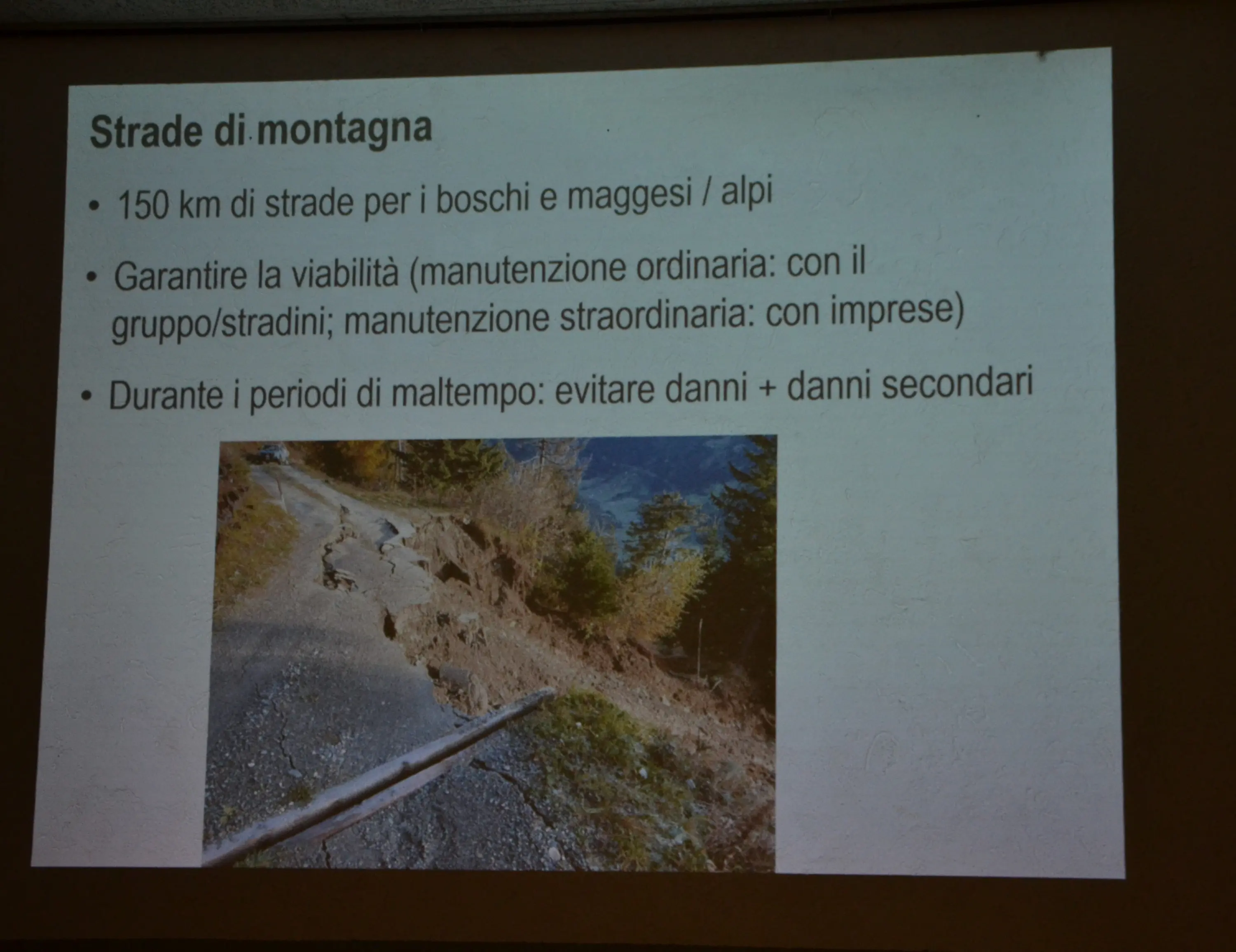Slide informativa sulle strade di montagna. Contiene un elenco di punti riguardanti la manutenzione stradale e la gestione dei danni, accompagnata da un'immagine di una strada danneggiata e dissestata.