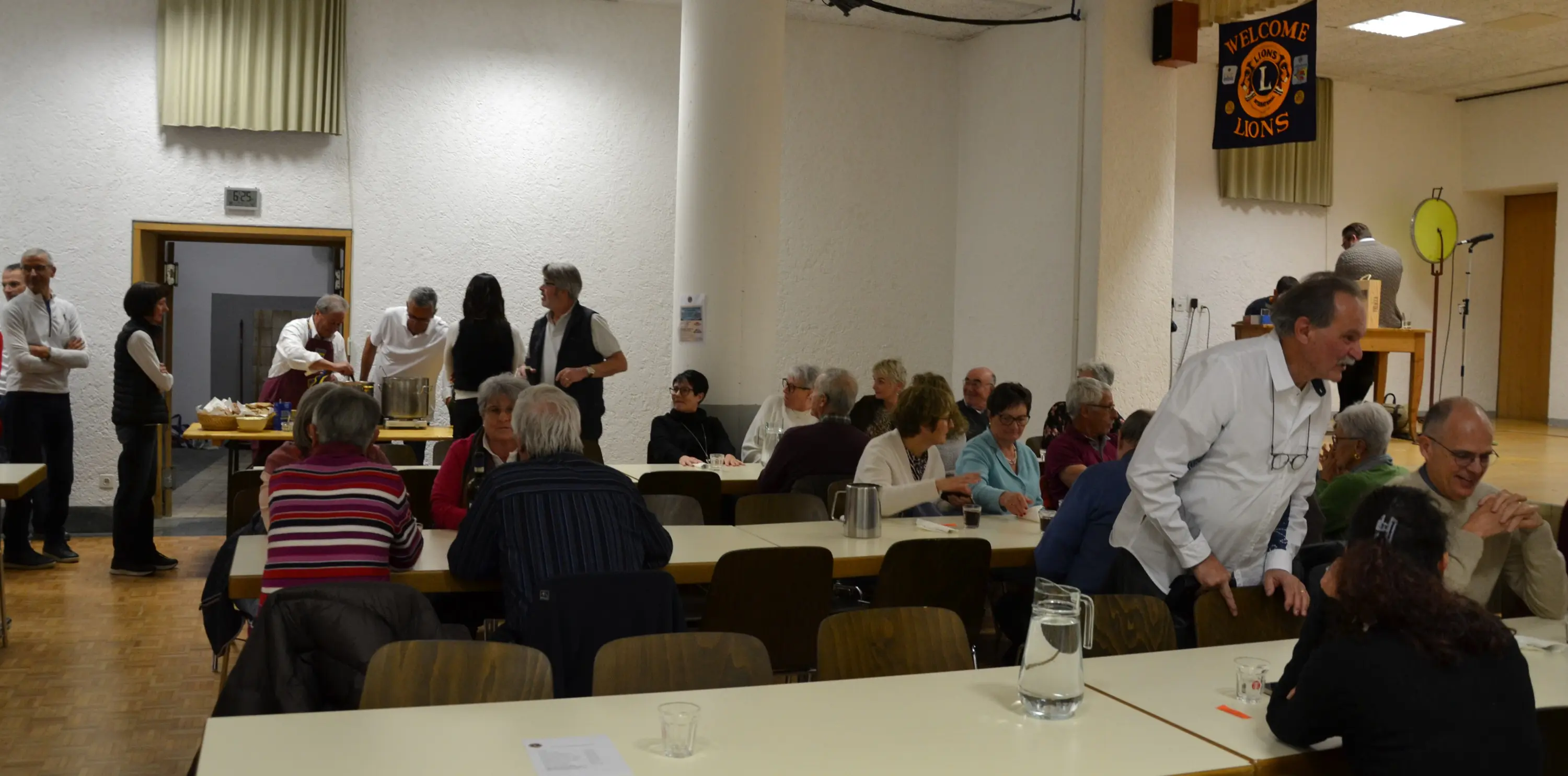 Una sala di comunità con persone sedute a lungo tavoli, mentre alcune persone servono cibo e bevande. In fondo, un uomo è in piedi accanto a un tavolo con un microfono. Un banner "Welcome Lions" è appeso alla parete.