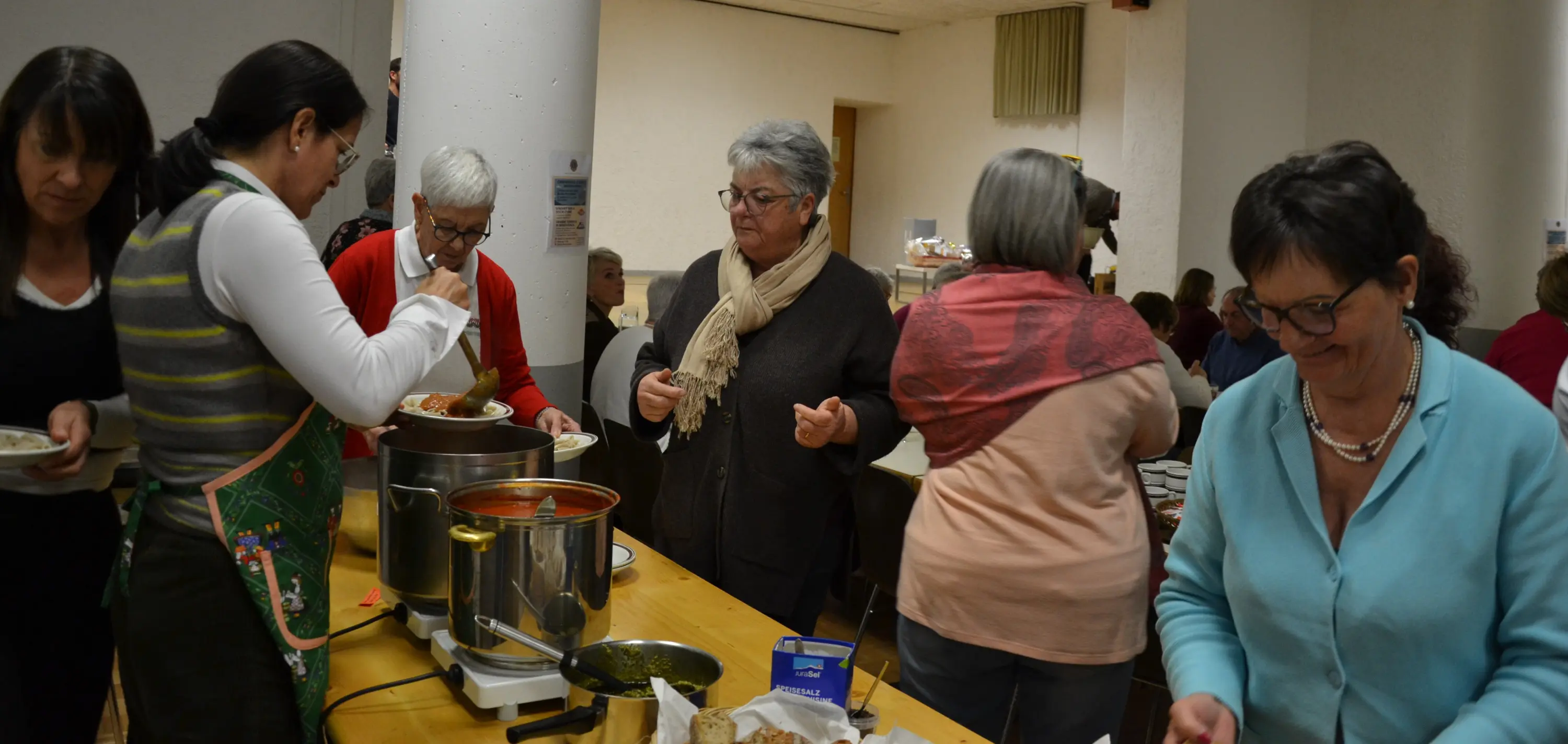 Un gruppo di donne è riunito in una cucina, dove alcune servono cibo da pentole in un tavolo. Si vede una varietà di piatti e ingredienti. C'è un'atmosfera conviviale e di collaborazione.
